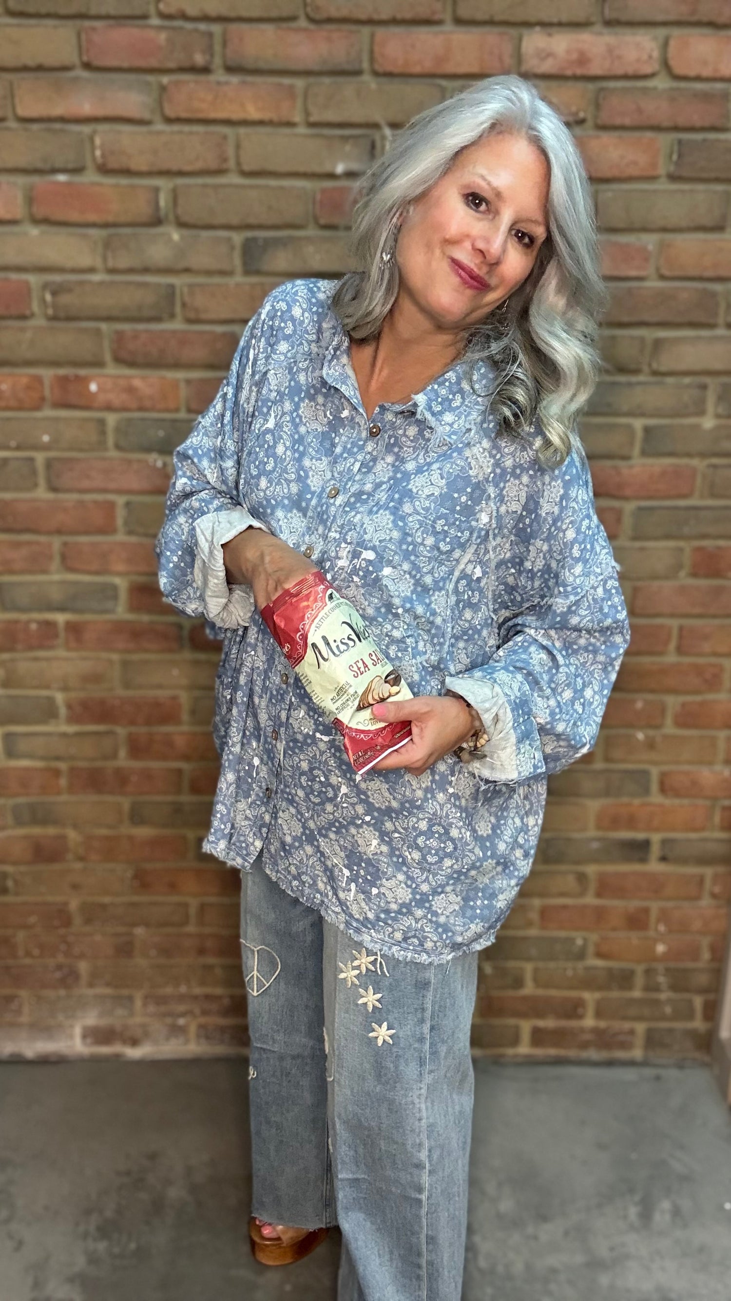 A woman with gray hair stands against a brick wall wearing a light blue and white floral print button-up shirt and light gray jeans, holding a red and white bag of chips.