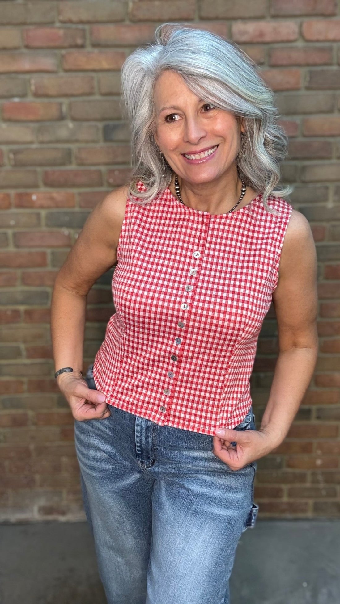 Woman wearing the Red Plaid Sleeveless Shirt  and blue jeans standing against a brick wall.