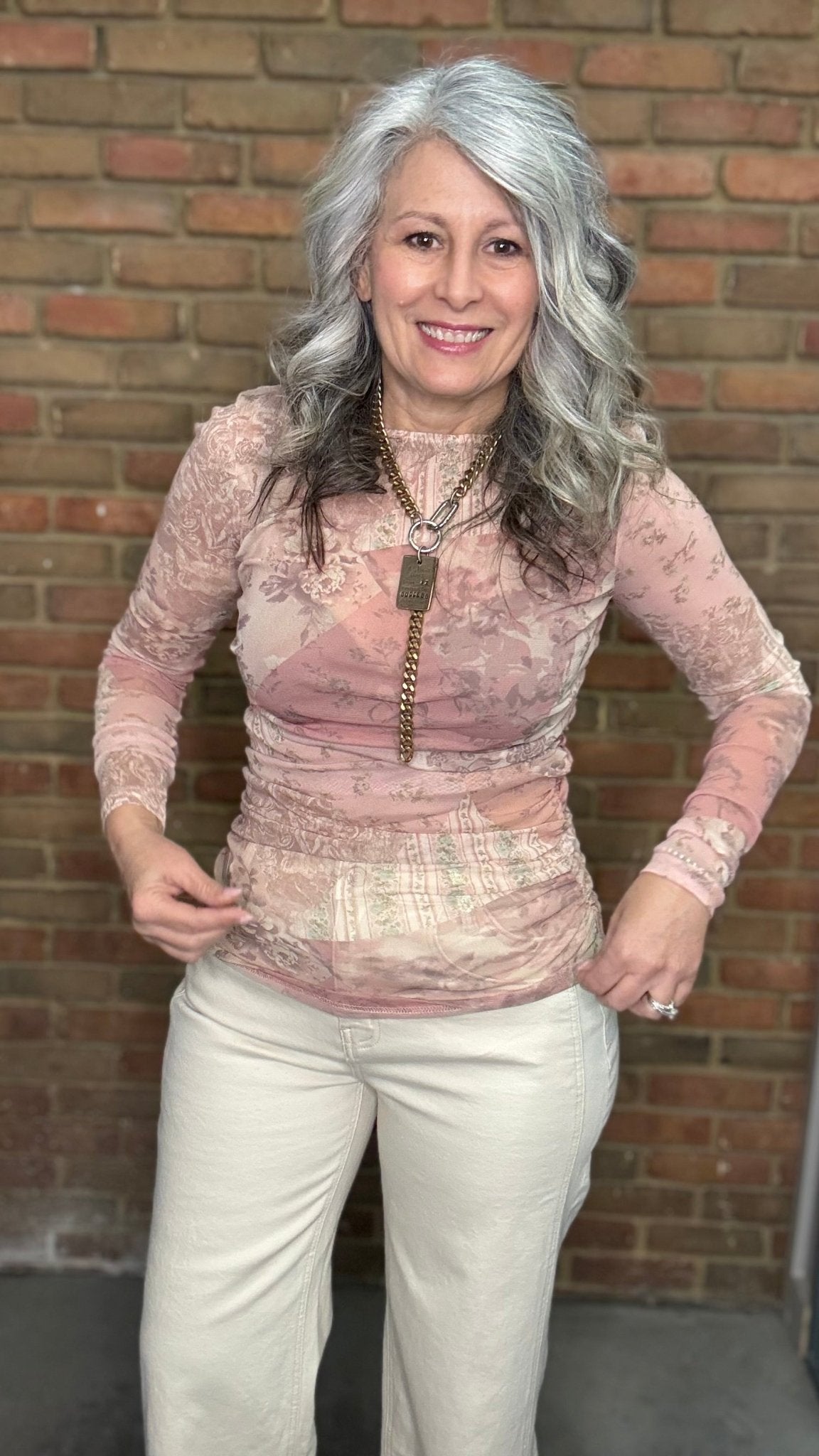 Woman wearing Mix Print Long Sleeve Mesh Top and white pants standing against a brick wall.