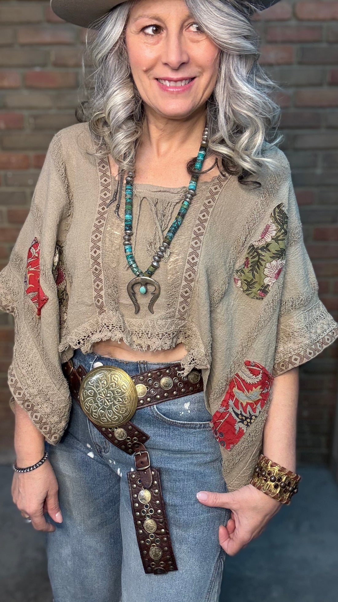 Woman wearing Garden Drift Top with a hat, jeans, and belt against a brick wall.