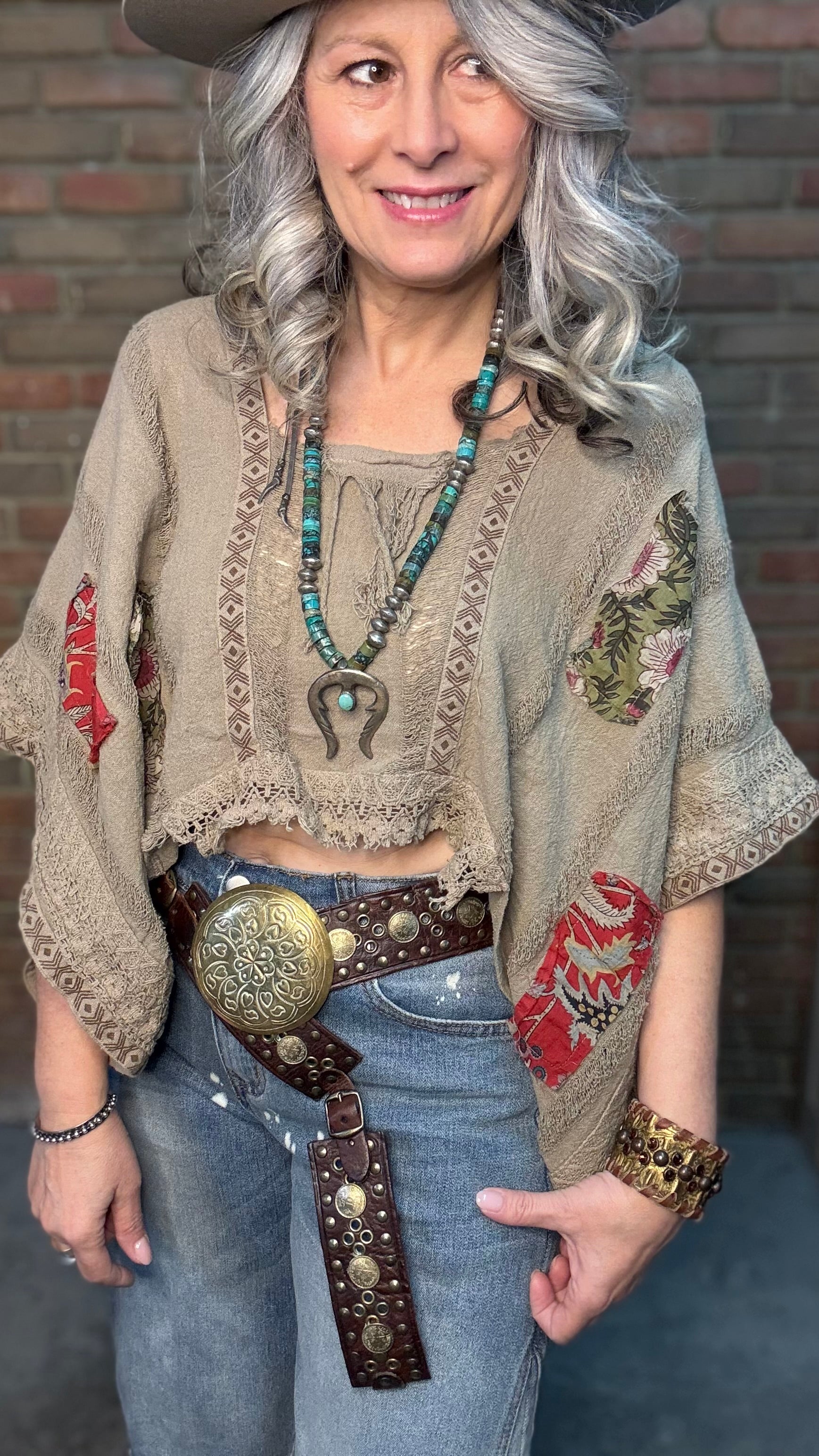 Woman wearing Garden Drift Top with a hat, jeans, and belt against a brick wall.
