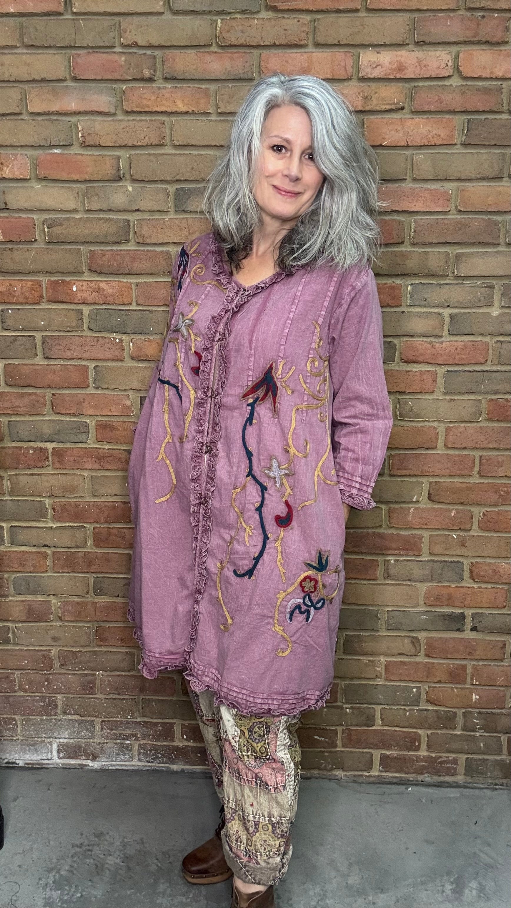 Woman wearing a purple embroidered jacket and patterned pants standing against a brick wall.