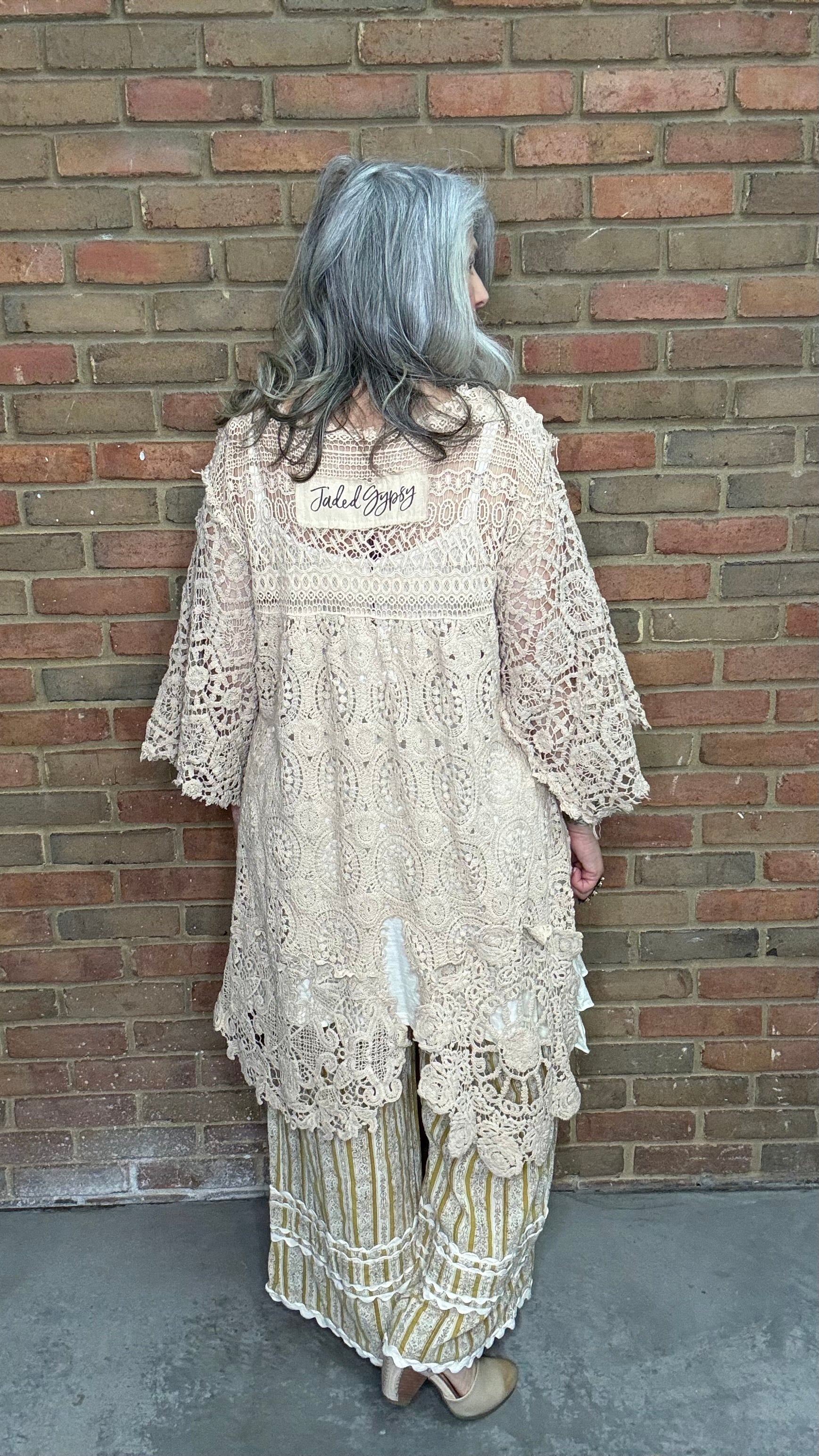 Woman wearing the crochet Forever Timeless Split Tails Jacket over a dress with striped pants standing against a brick wall.