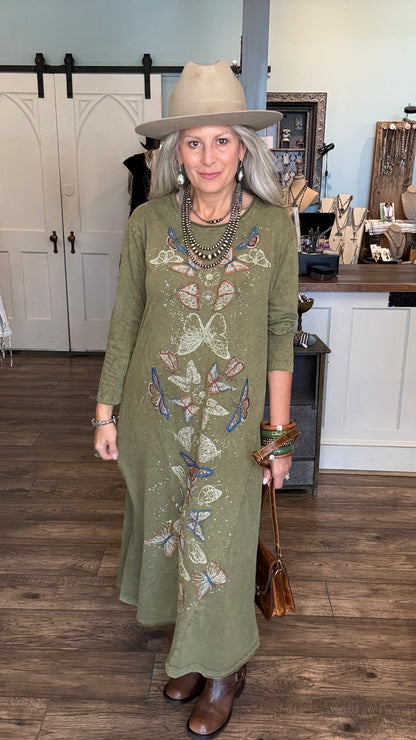 A woman wearing a sage green long-sleeve maxi dress with butterfly embroidery, a beige wide-brim hat, and brown boots, standing in a boutique setting.