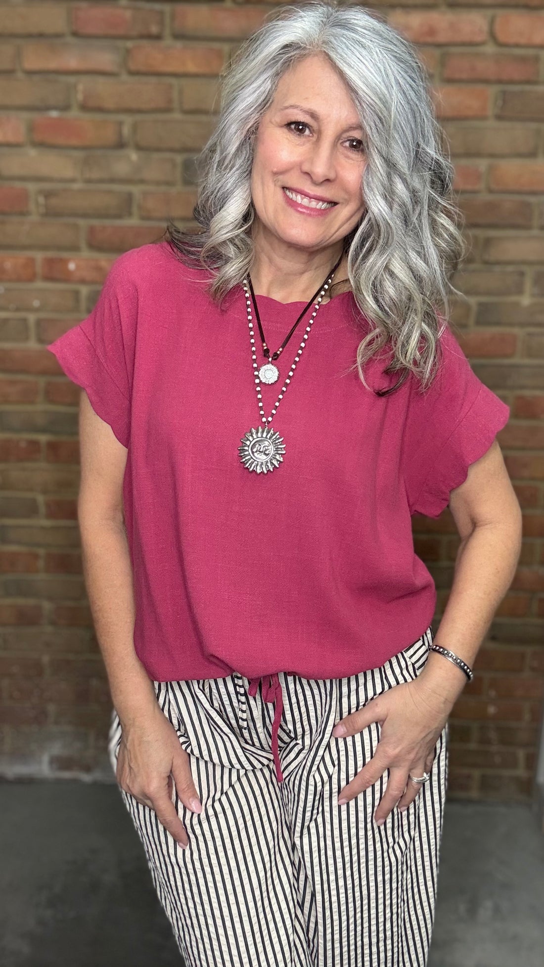 Woman wearing Scalloped Sleeve Tie Hem Top - Berry and striped pants standing against a brick wall.