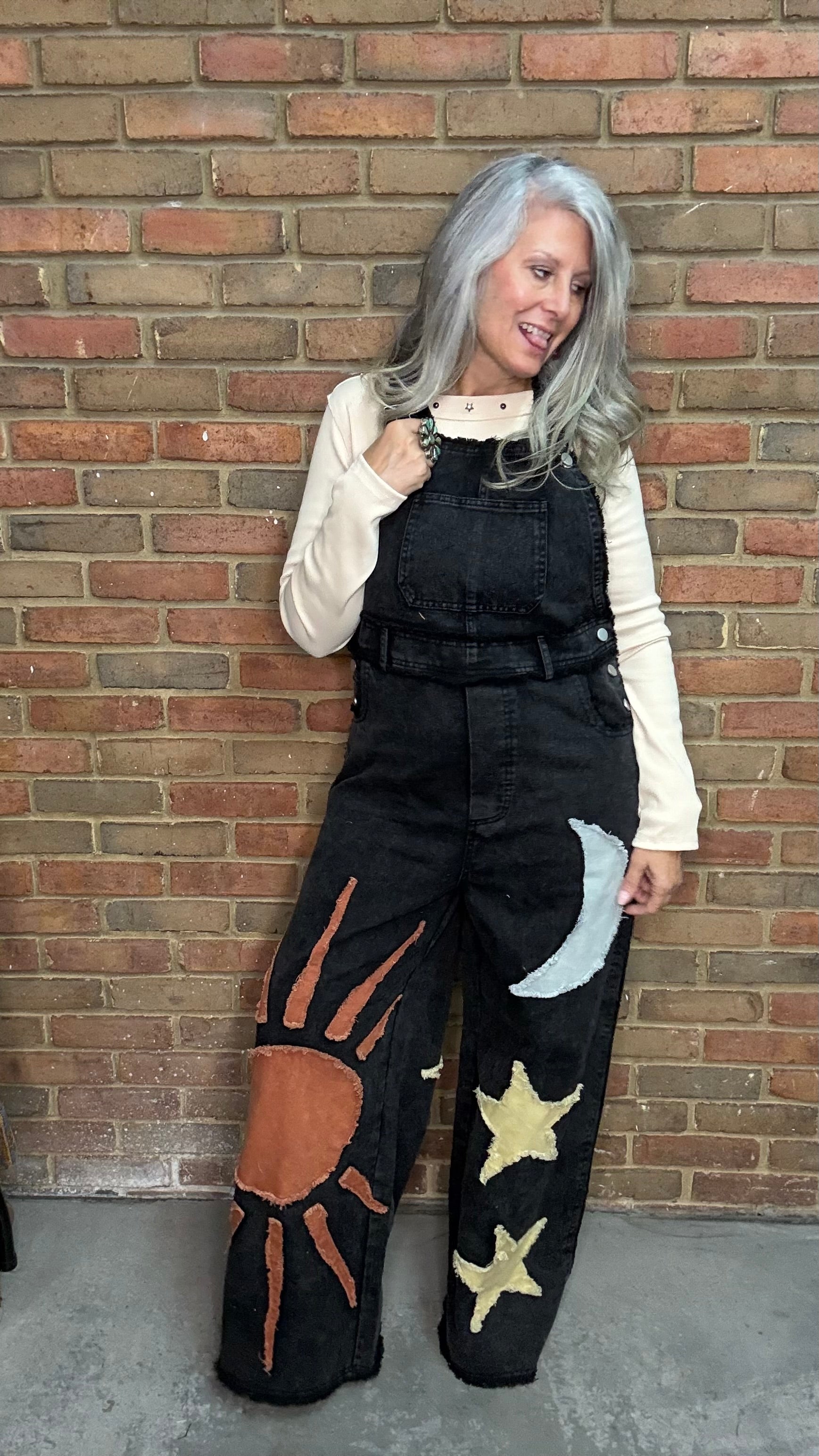 A woman with silver hair stands against a red brick wall wearing black denim overalls with celestial patches (sun, moon, stars) over a cream long-sleeve top.
