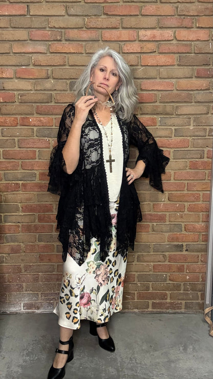 Woman in a black kimono-style jacket over a floral dress against a brick wall.