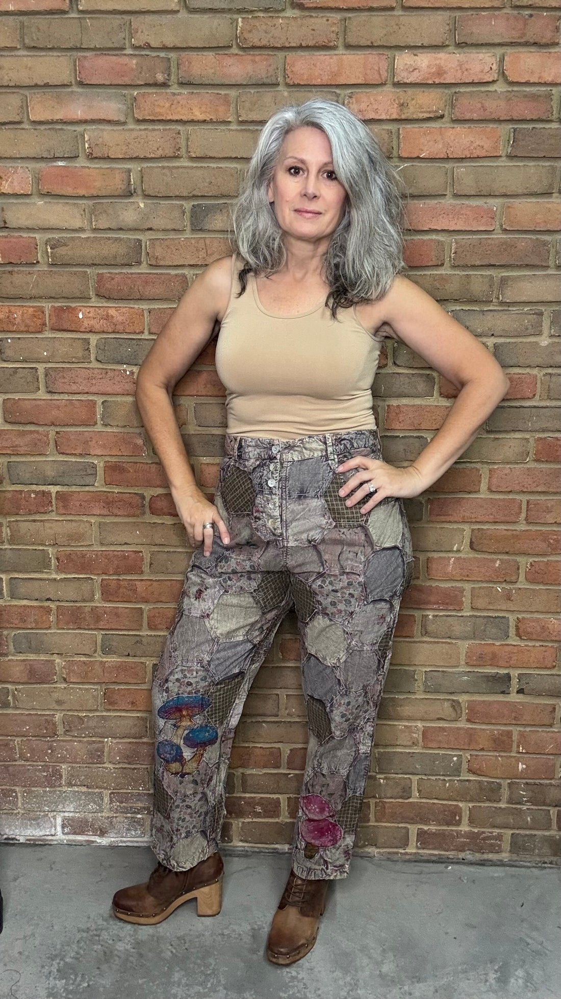 Person wearing a beige tank top and patchwork pants against a brick wall.