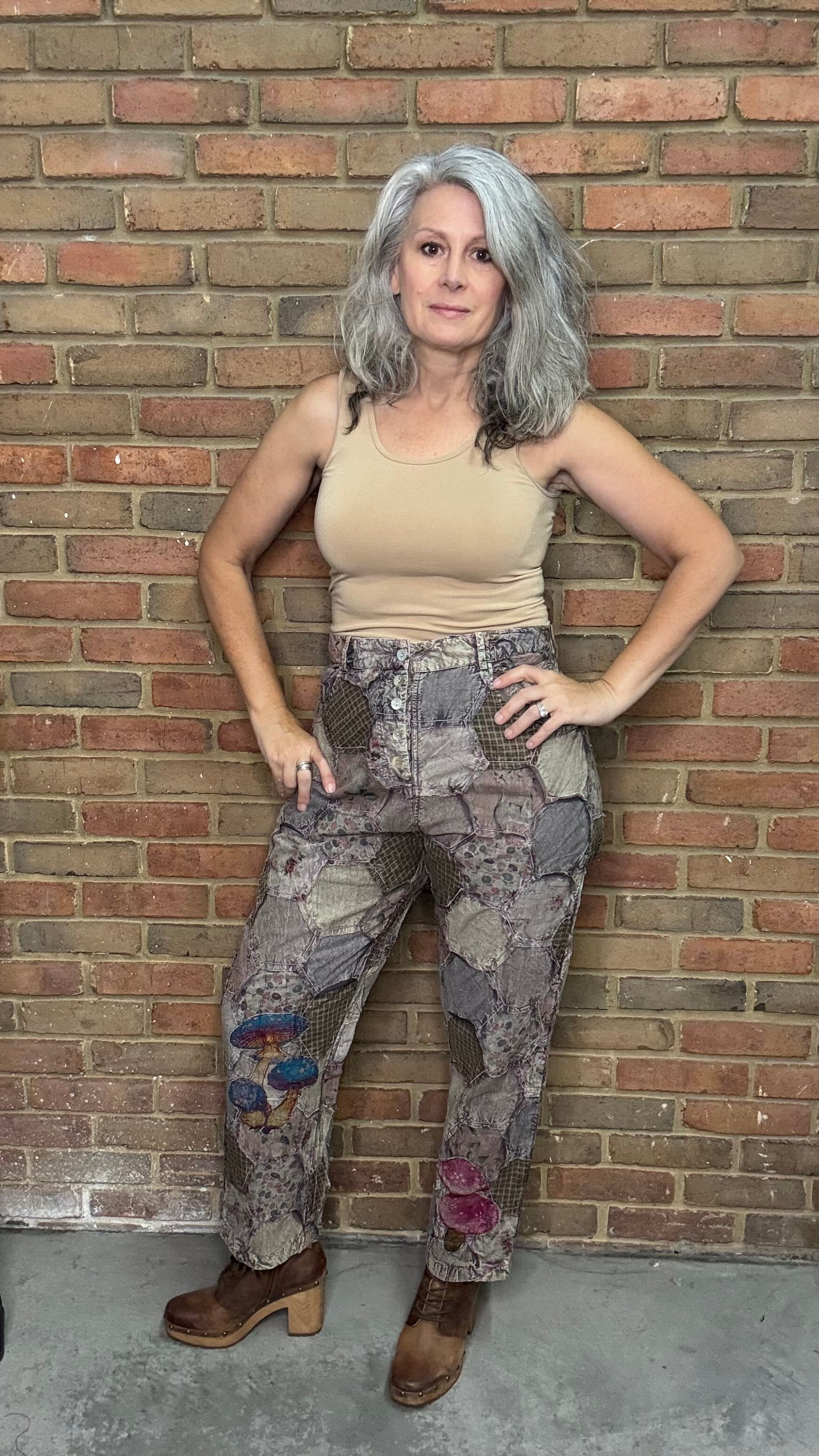 Person wearing a beige tank top and patchwork pants against a brick wall.