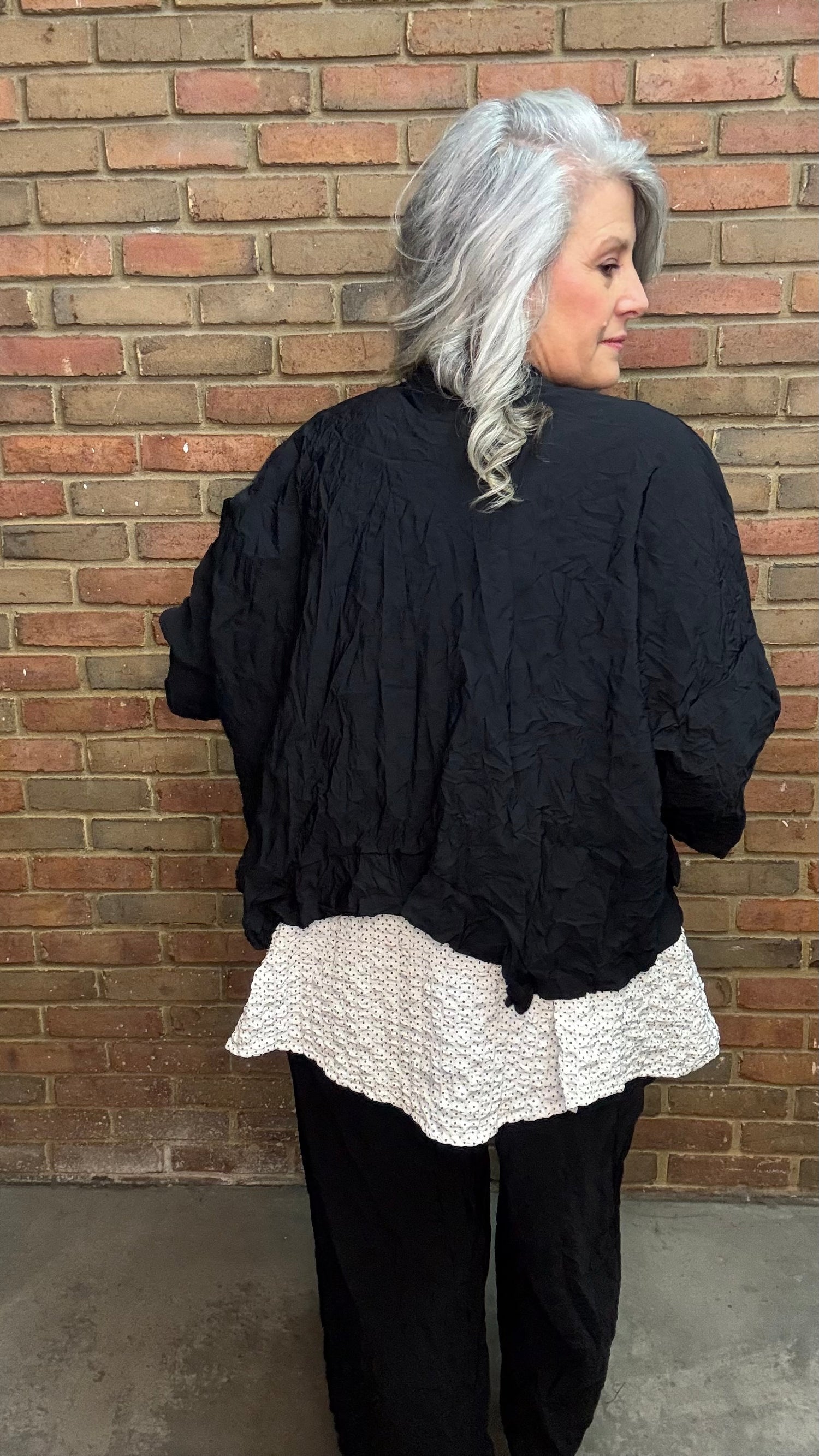 Woman standing against a brick wall wearing Francesca Jacket - Black, white top, and black pants.