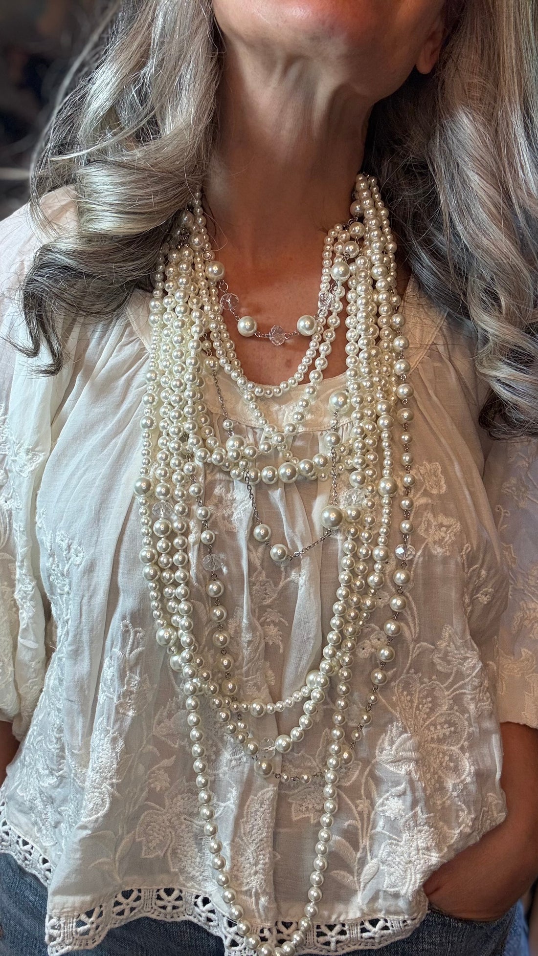 A close-up of a woman wearing a layered necklace set with multiple strands of cream faux pearls styled over a white lace blouse.