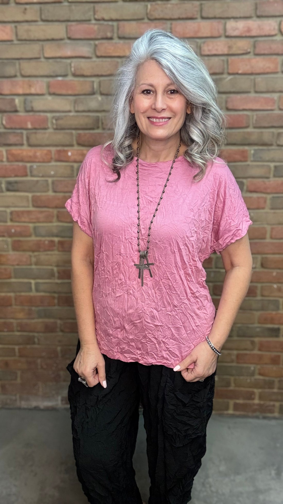 Woman with gray hair wearing Rose Dolman Sleeve Top and black pants standing in front of a brick wall.
