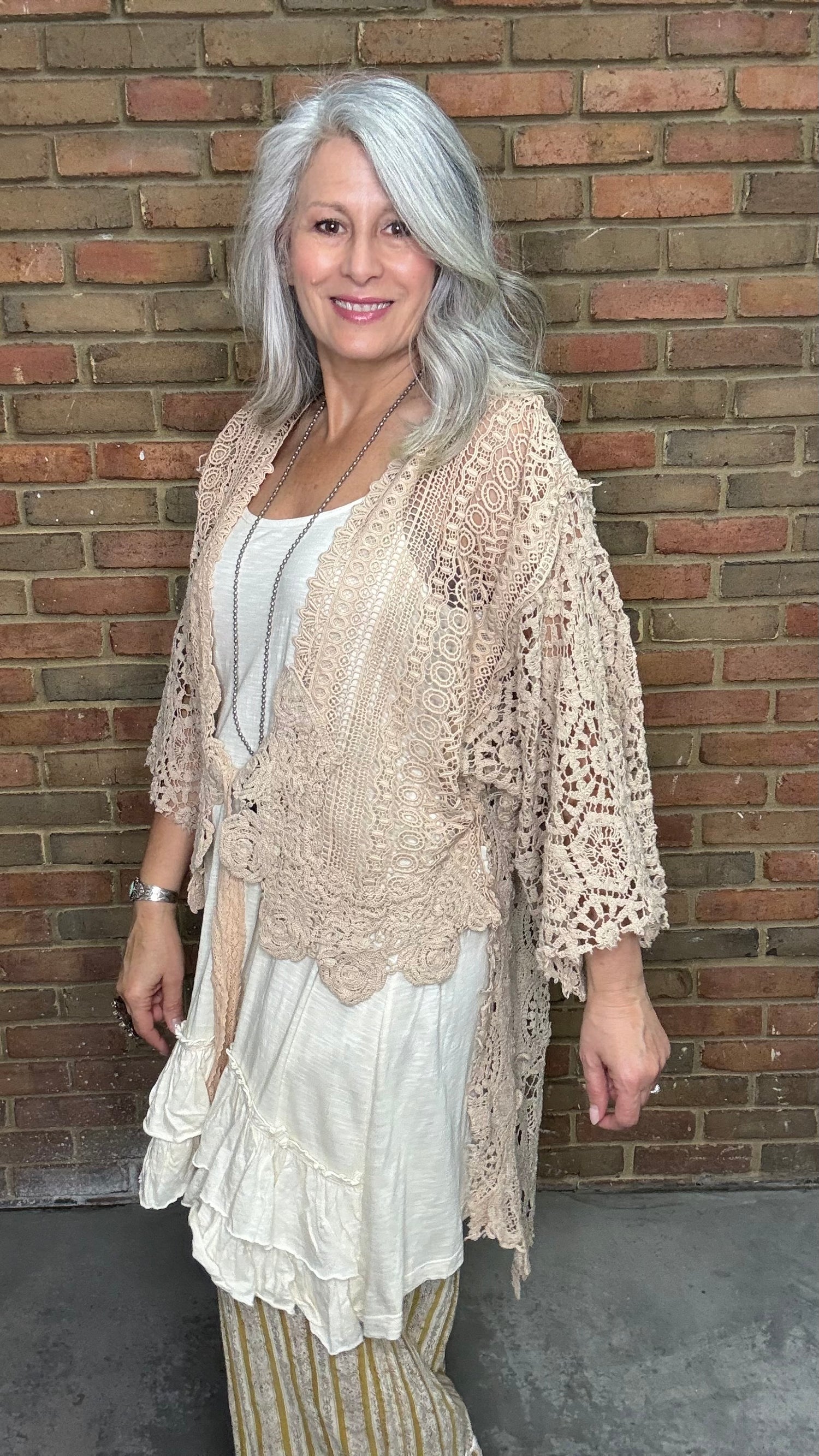 Woman wearing the crochet Forever Timeless Split Tails Jacket over a dress with striped pants standing against a brick wall.