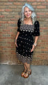 A woman with silver hair wearing a black short-sleeve dress with white embroidered Celtic knot patterns and star motifs, standing against a brick wall.