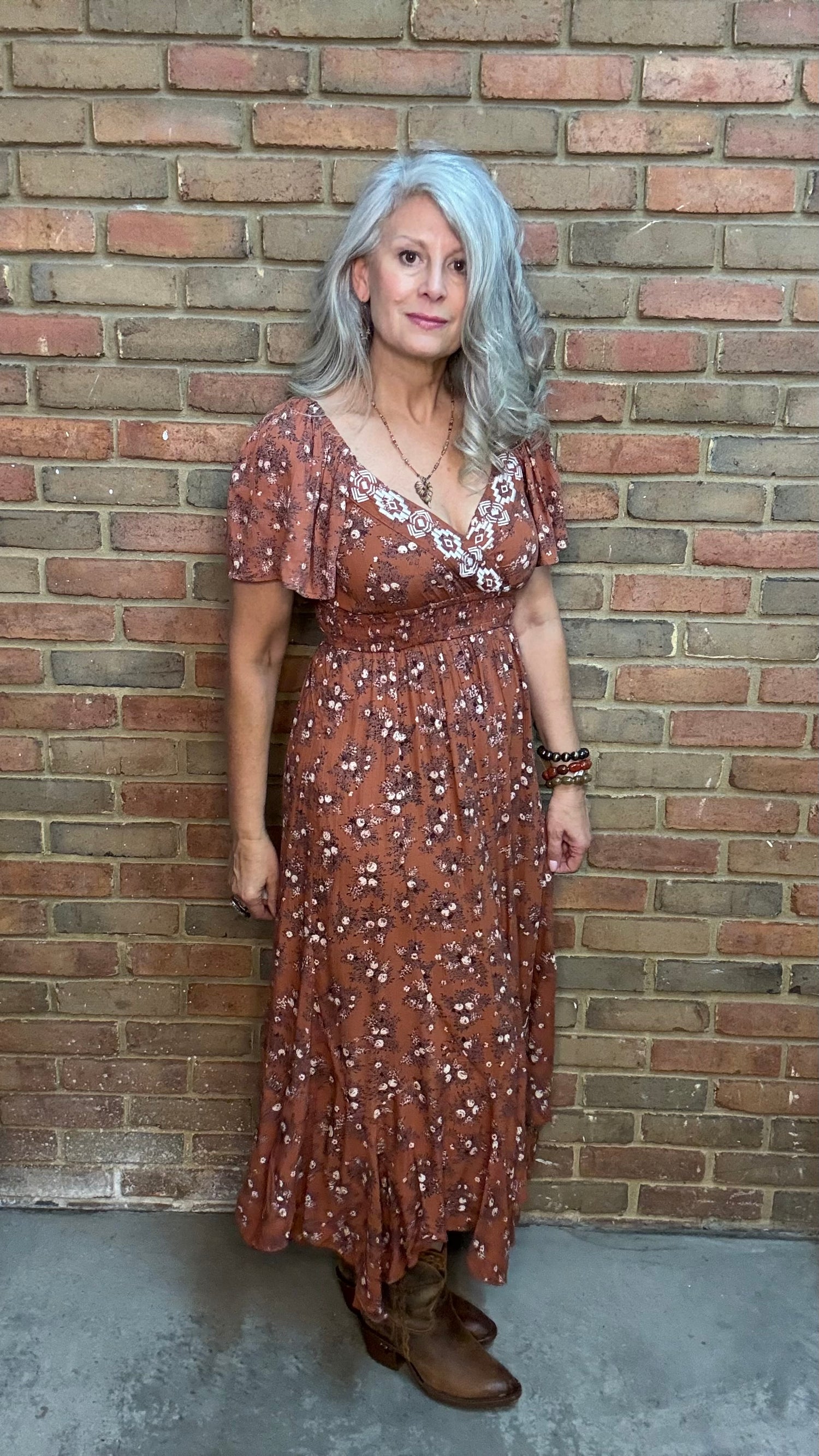 A woman with gray hair stands against a brick wall wearing a rust-brown bohemian-style midi dress with white floral prints, the "Short Sleeve Embroidered Midi Dress" and brown cowboy boots.