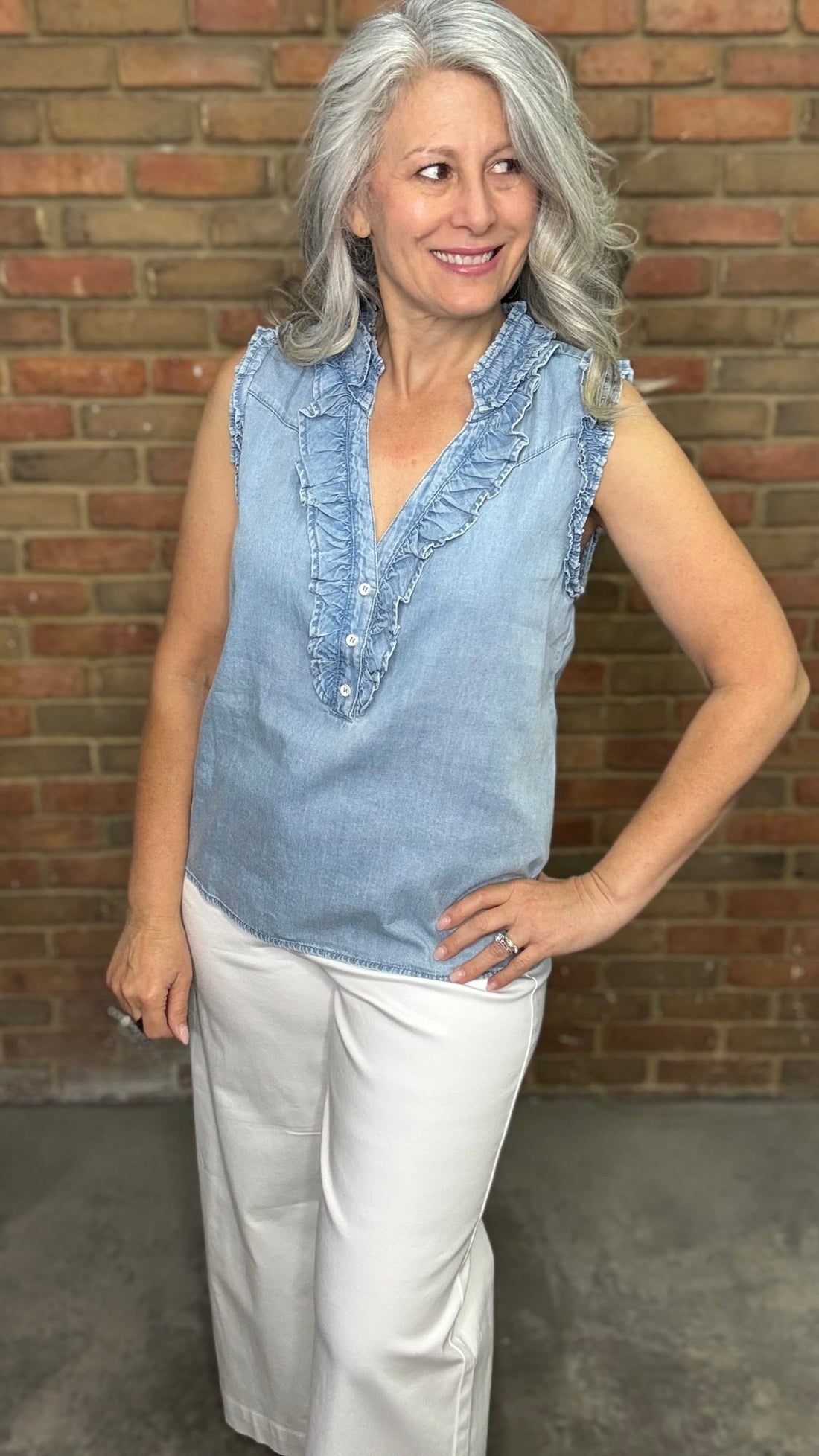 Woman wearing the Ruffle Sleeveless V-Neck Top, a light blue sleeveless top with ruffled details and white pants against a brick wall.