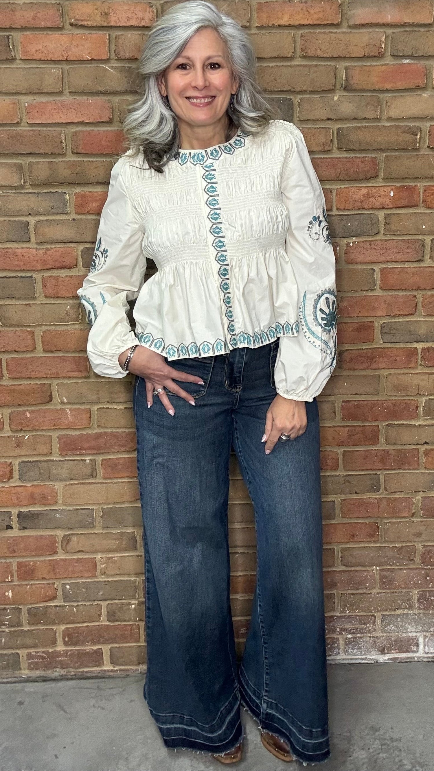Woman wearing the Embroideried Smocked Peasant Top and Judy Blue Palazzo jeans against a brick wall.