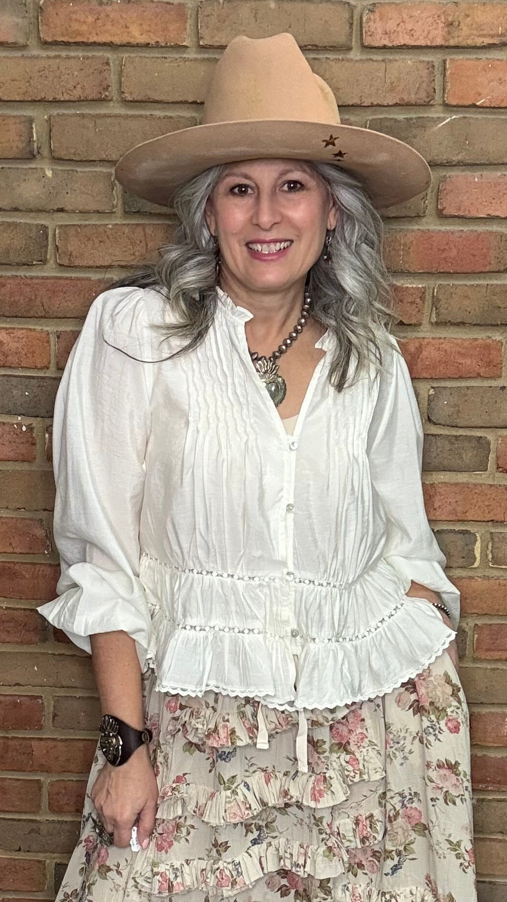 Woman in a White Pin-Tuck Lace Embellished shirt, blooming romance skirt, and Kate Harness Frye boots standing against a brick wall.