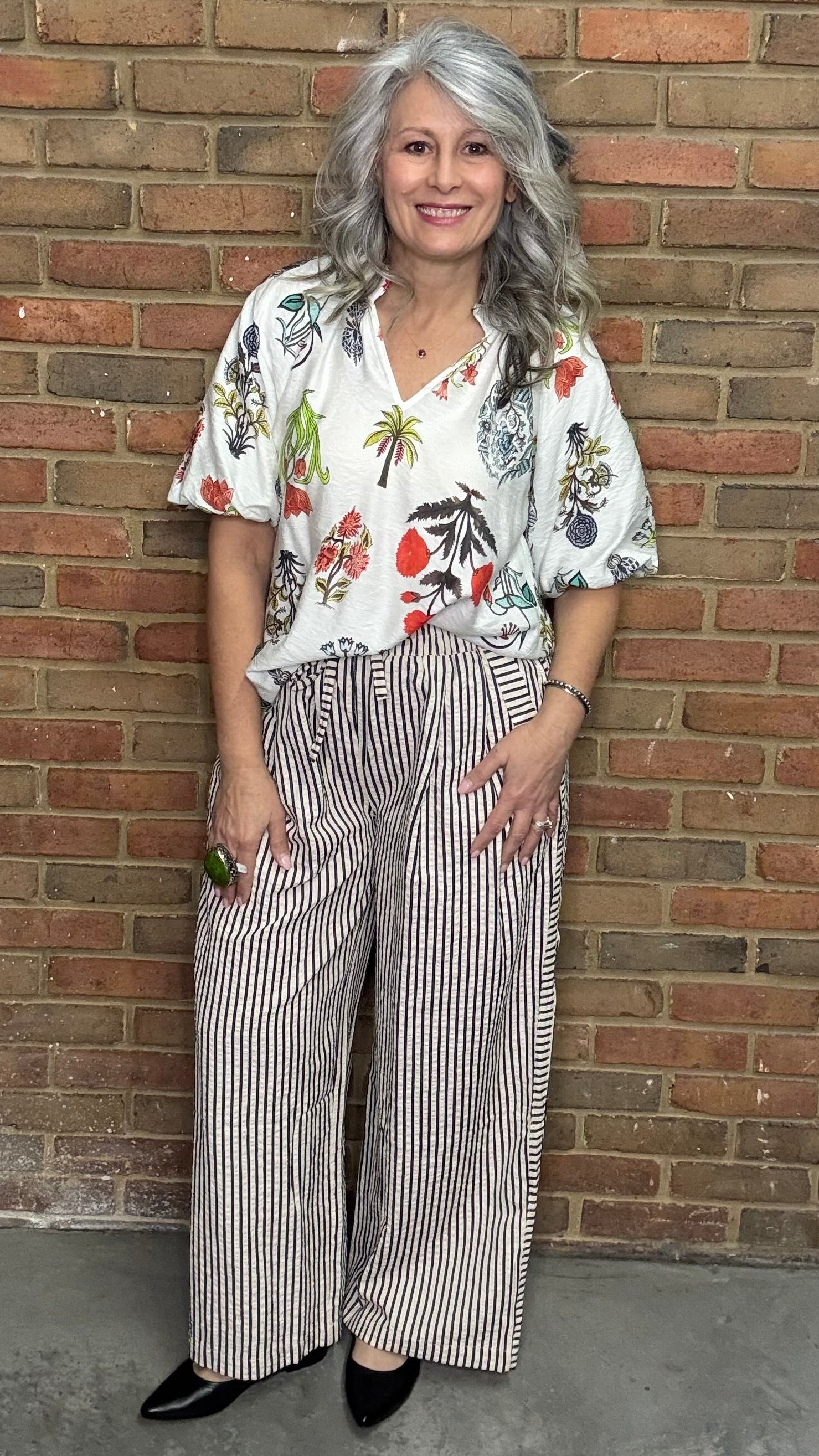 Woman in a floral blouse and Latte Wide Leg Pull On Pant standing against a brick wall.