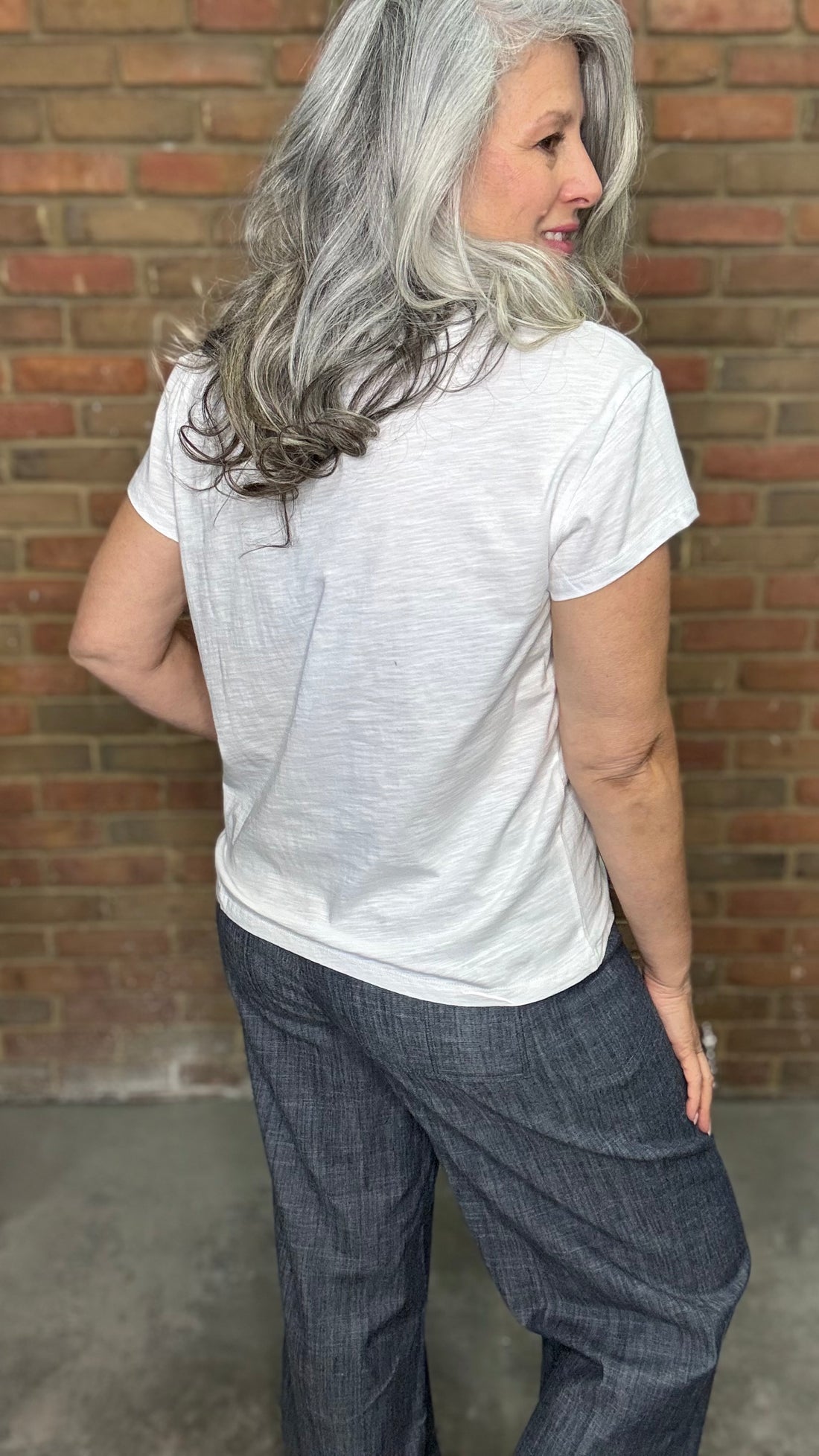 Woman wearing White Drop Shoulder V-Neck Top and Indigo Linen Button Front Pants standing against a brick wall.