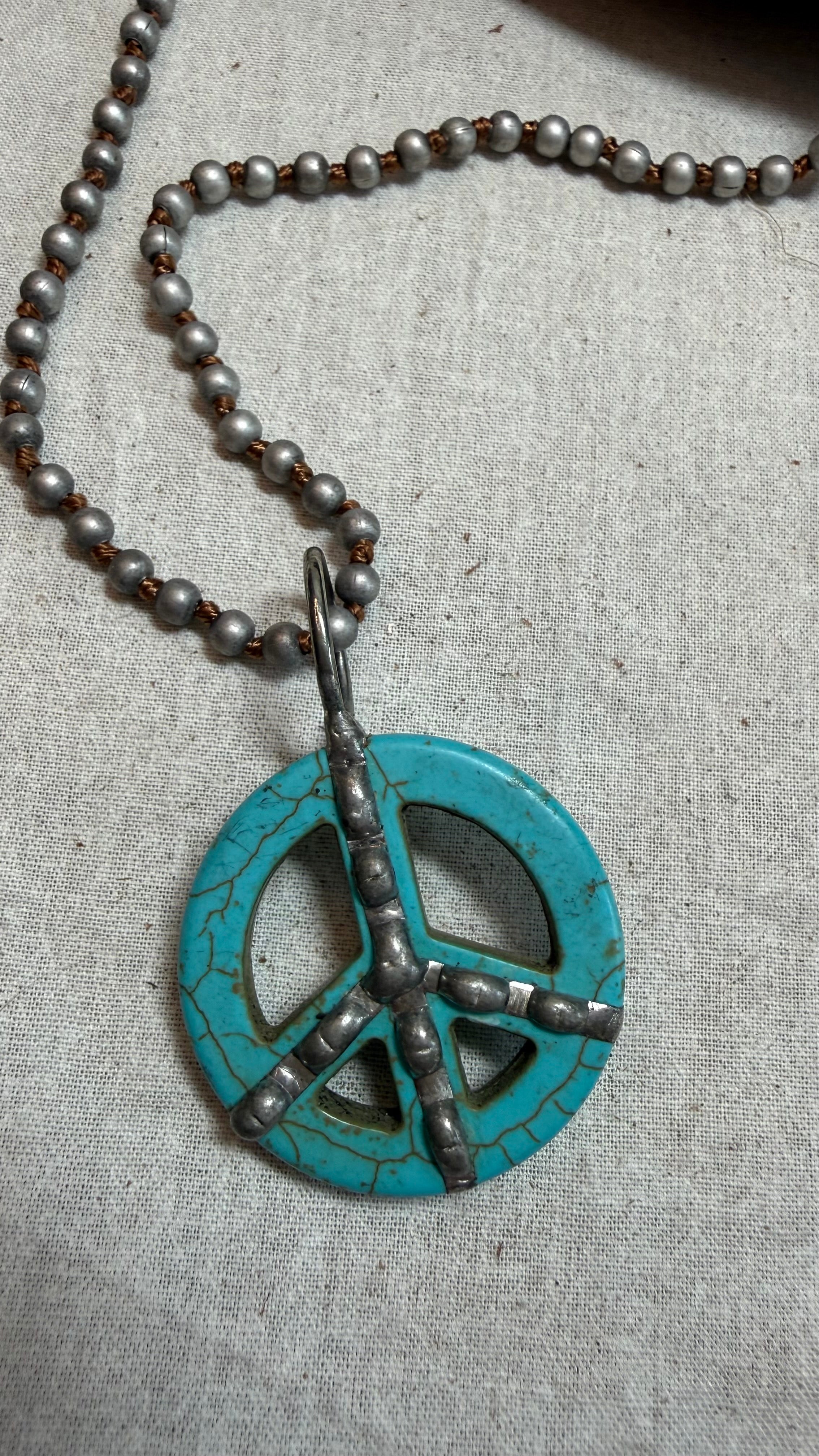 A turquoise and silver peace sign pendant necklace on a matching beaded chain, displayed on a textured beige linen fabric background.