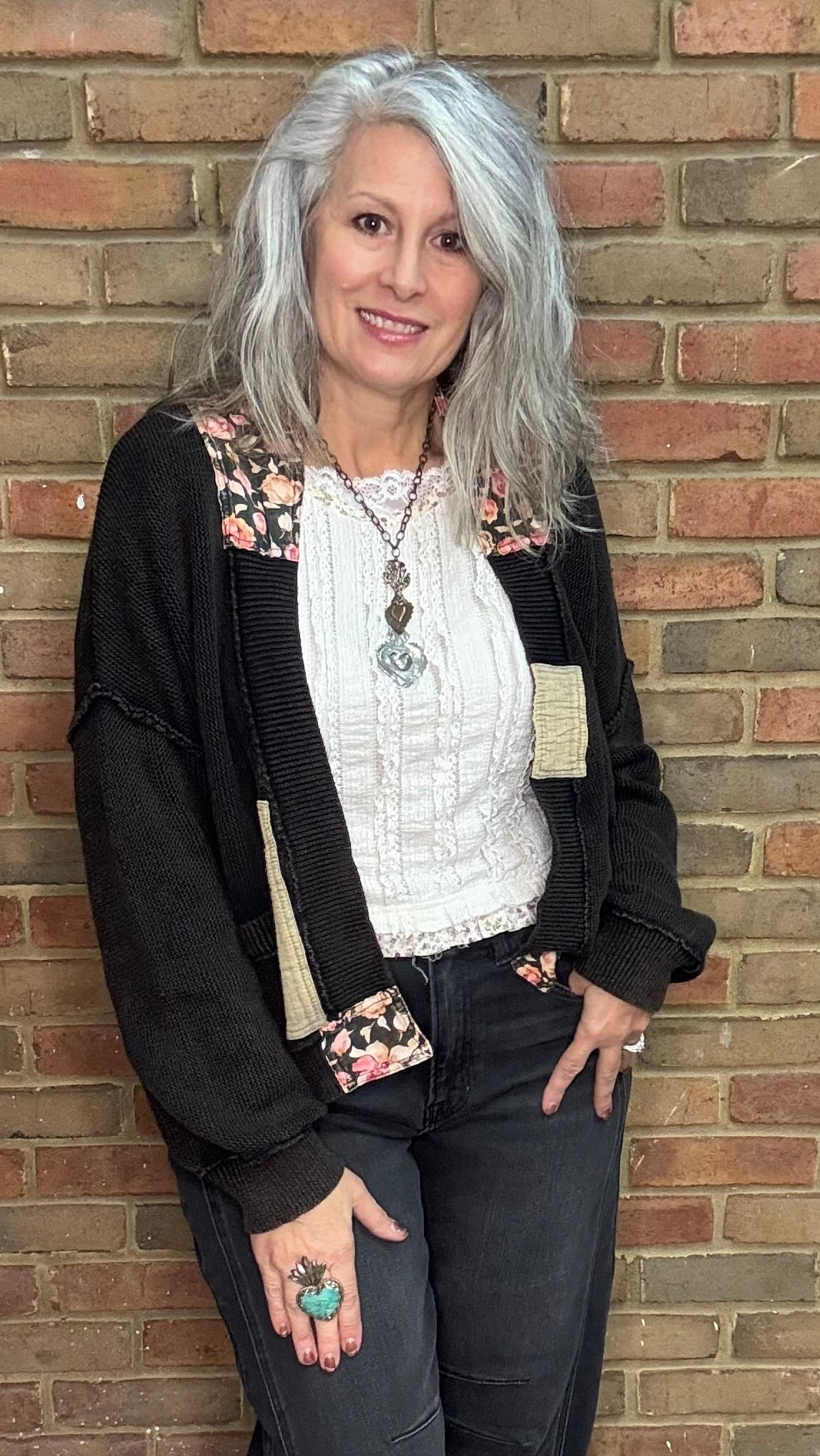 Woman standing against a brick wall wearing a black cardigan, white top, and dark pants.