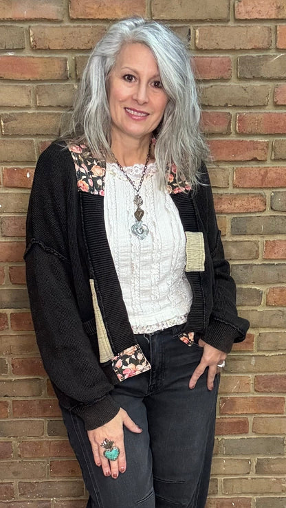 Woman standing against a brick wall wearing a black cardigan, white top, and dark pants.