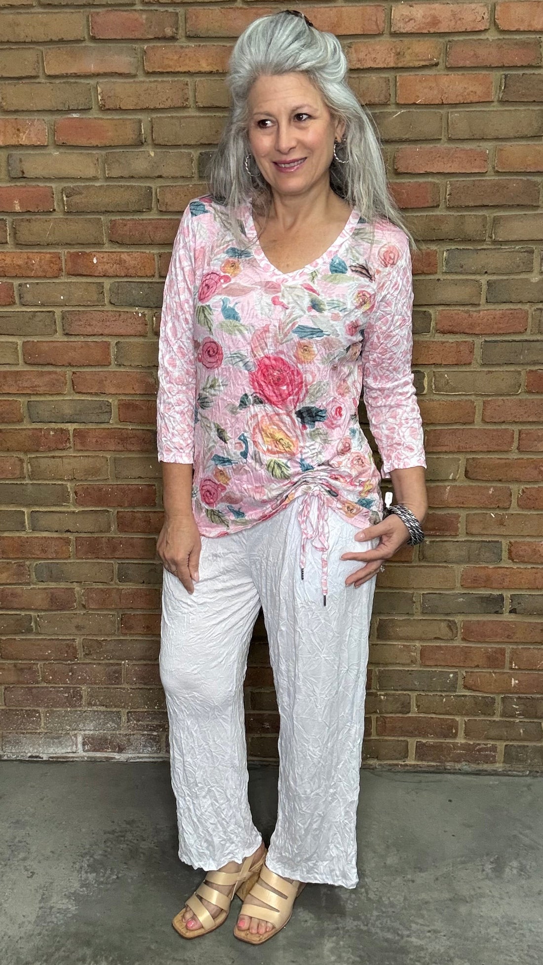 Woman standing against a brick wall wearing the Pink Floral Cinch Side Top and white pants.