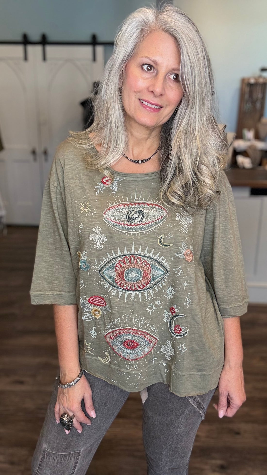 A woman with long gray hair wearing a sage green graphic t-shirt with embroidered eyes and celestial designs, paired with gray jeans, standing in a home interior setting.