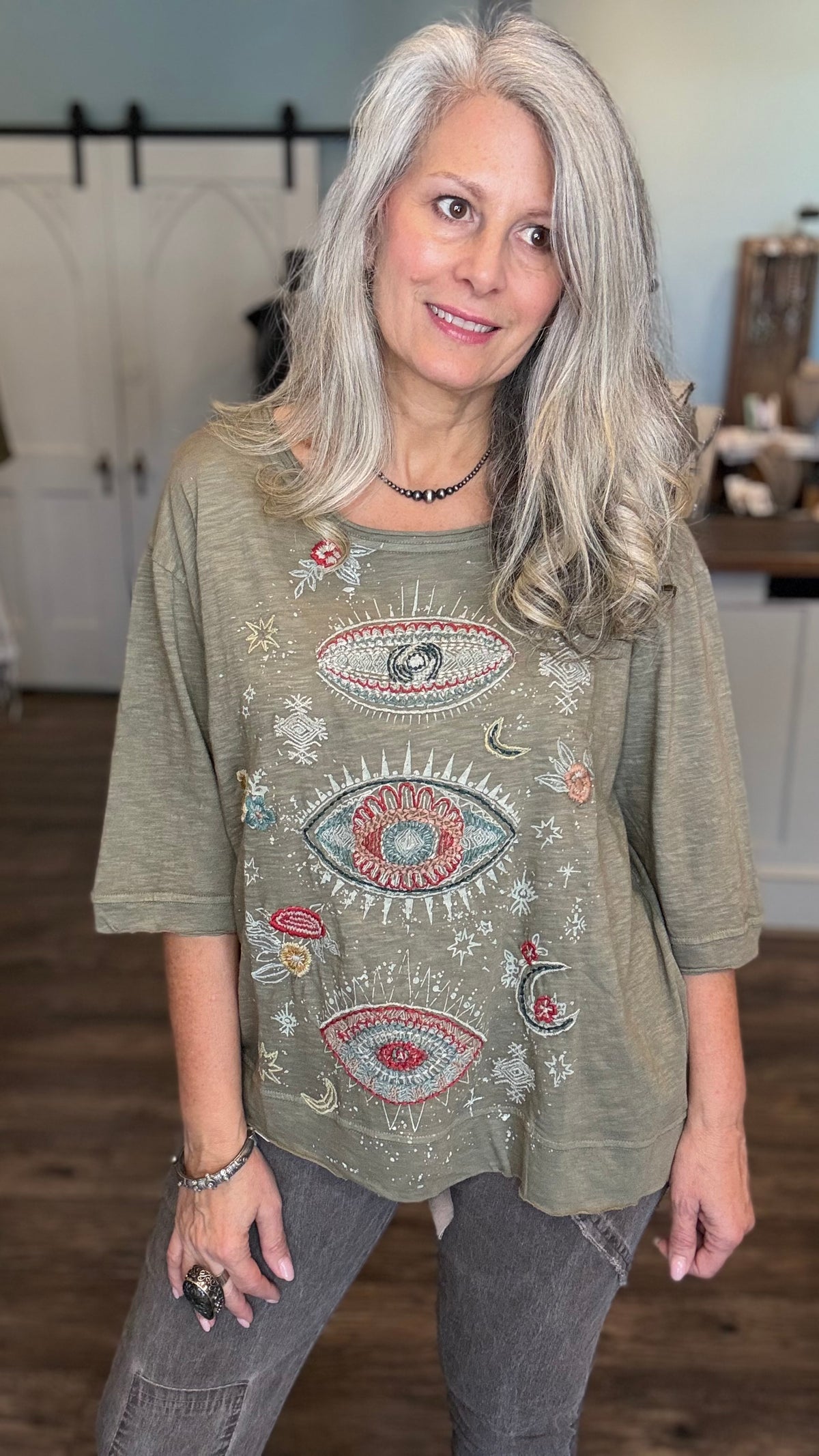 A woman with long gray hair wearing a sage green graphic t-shirt with embroidered eyes and celestial designs, paired with gray jeans, standing in a home interior setting.