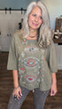 A woman with long gray hair wearing a sage green graphic t-shirt with embroidered eyes and celestial designs, paired with gray jeans, standing in a home interior setting.