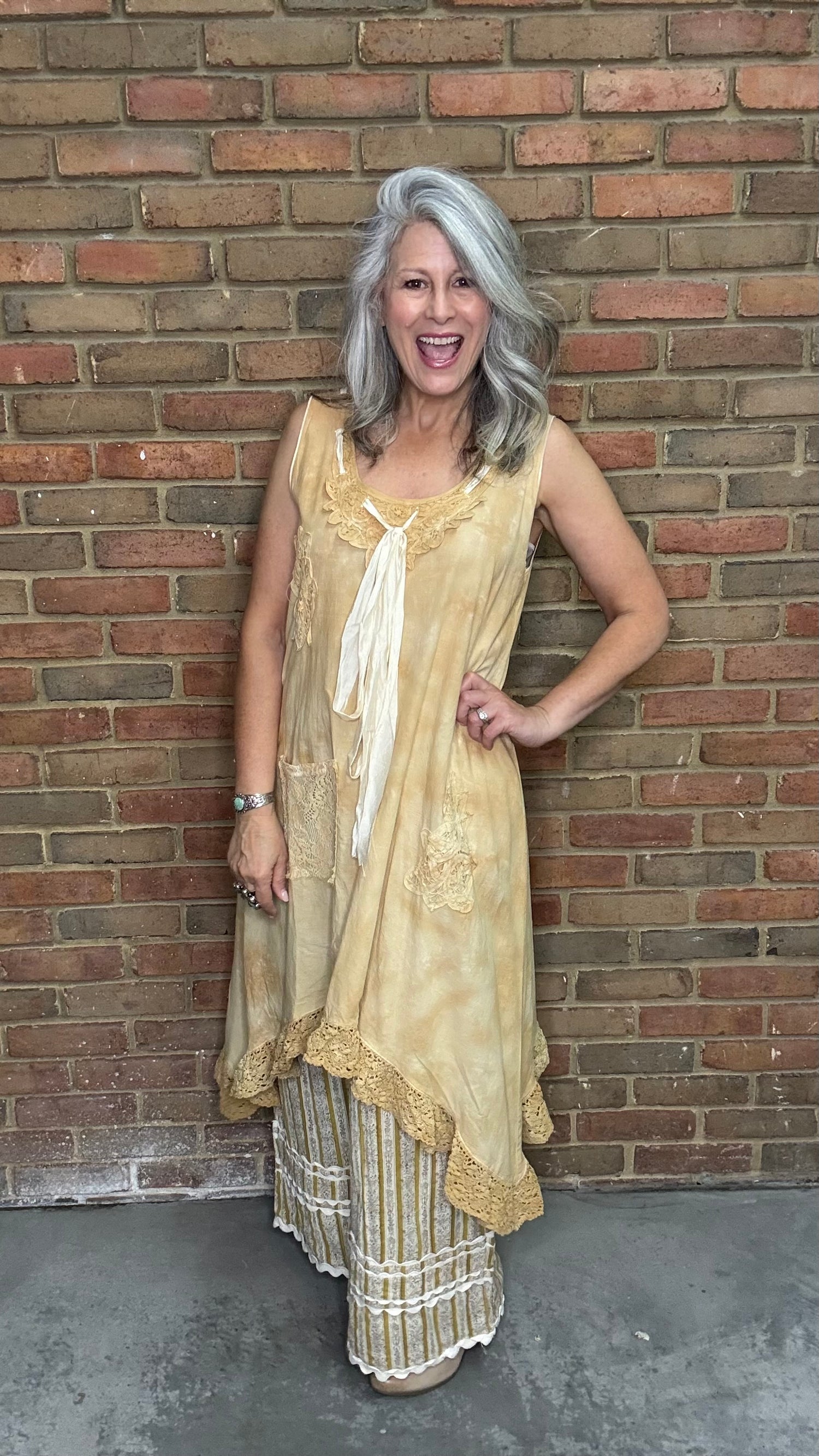 Woman wearing the Wandering Spirit Dress a tea stained tank dress with striped pants standing in front of a brick wall.