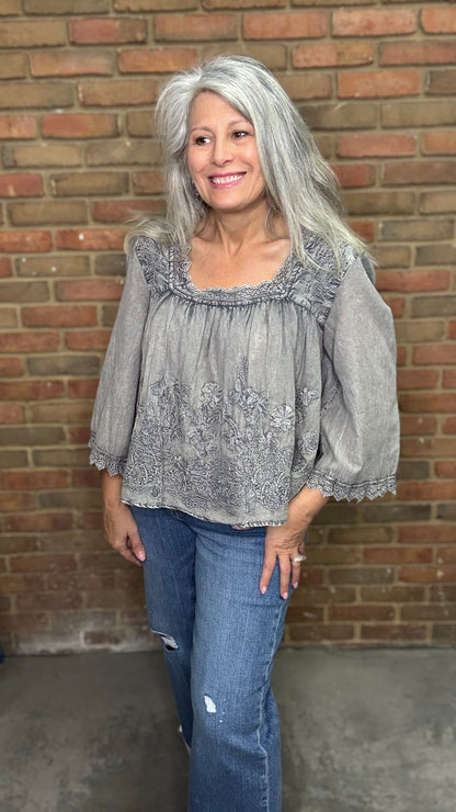 Woman with gray hair wearing the Vintage Washed Floral Lace Top a  light gray blouse and blue jeans standing against a brick wall.