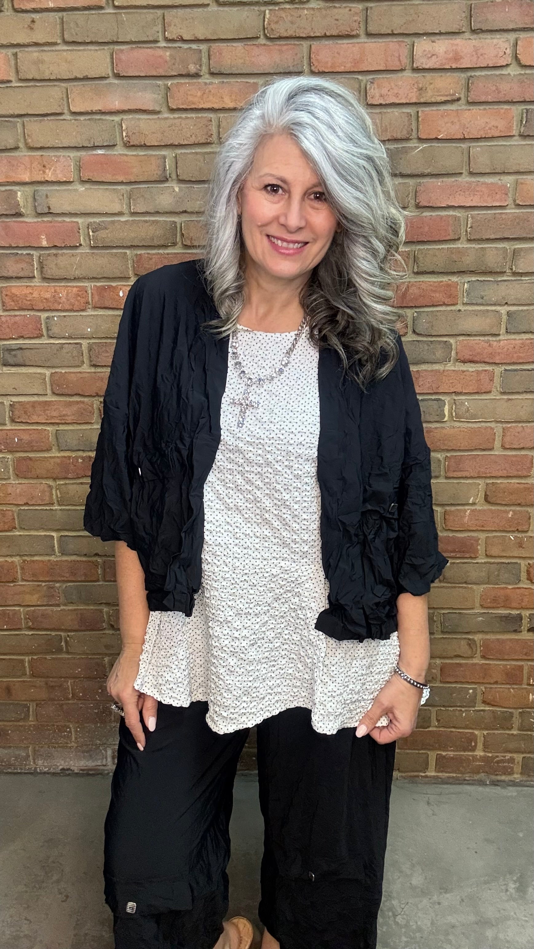 Woman standing against a brick wall wearing Francesca Jacket - Black, white top, and black pants.