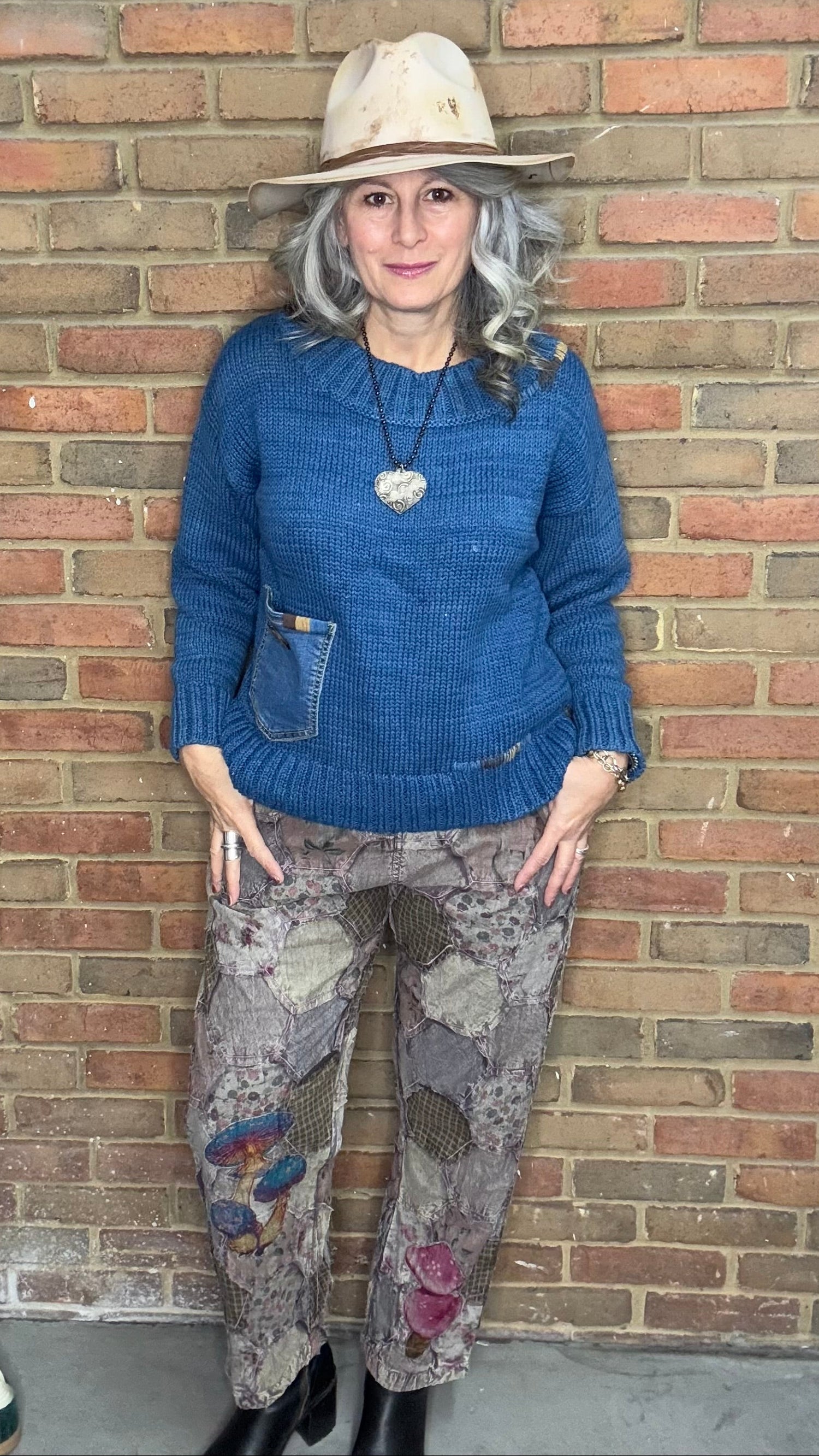 Person wearing a blue sweater and patterned pants standing against a brick wall.