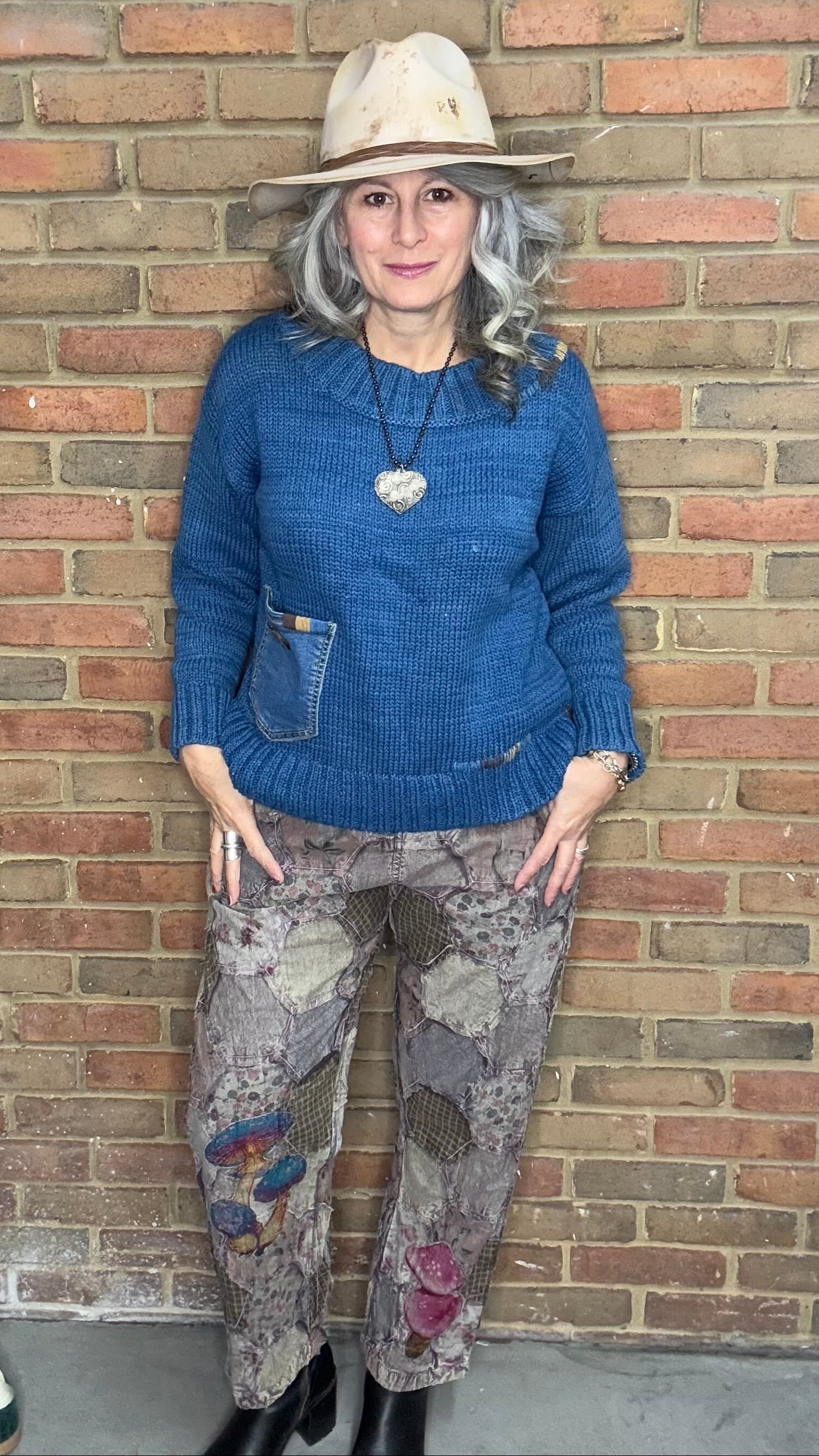 Person wearing a blue sweater and patterned pants standing against a brick wall.