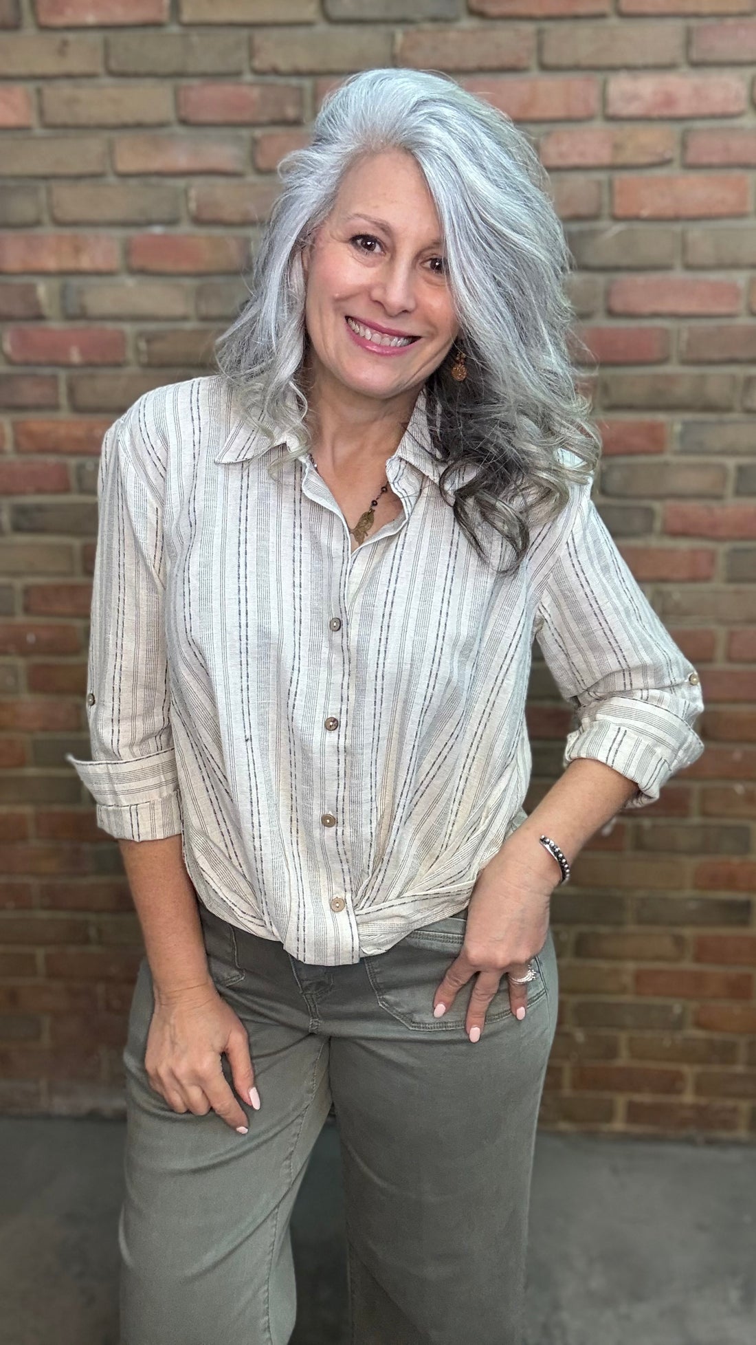 Woman wearing Yarn Dye Stripe Twist Shirt with pants against brick wall.