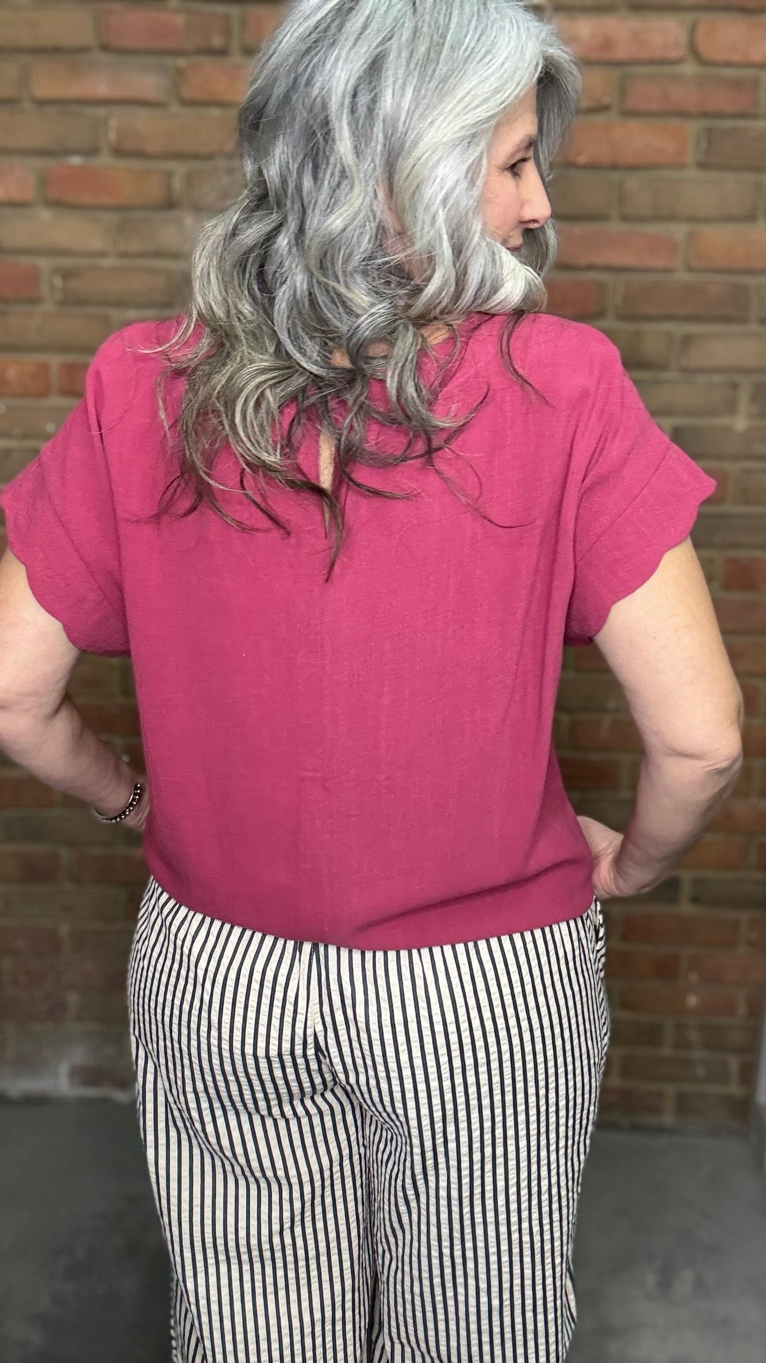 Woman wearing Scalloped Sleeve Tie Hem Top - Berry and striped pants standing against a brick wall.