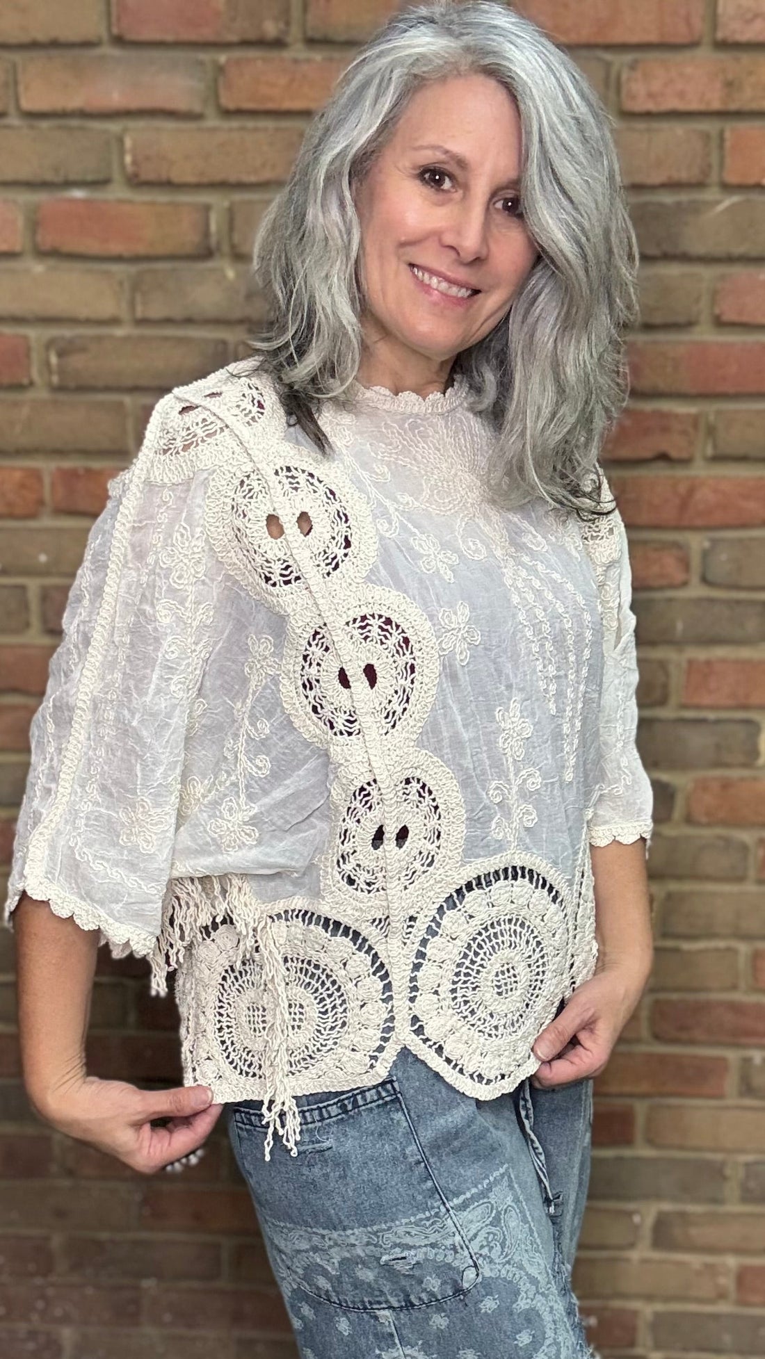 Woman wearing a white lace top and blue jeans standing against a brick wall.