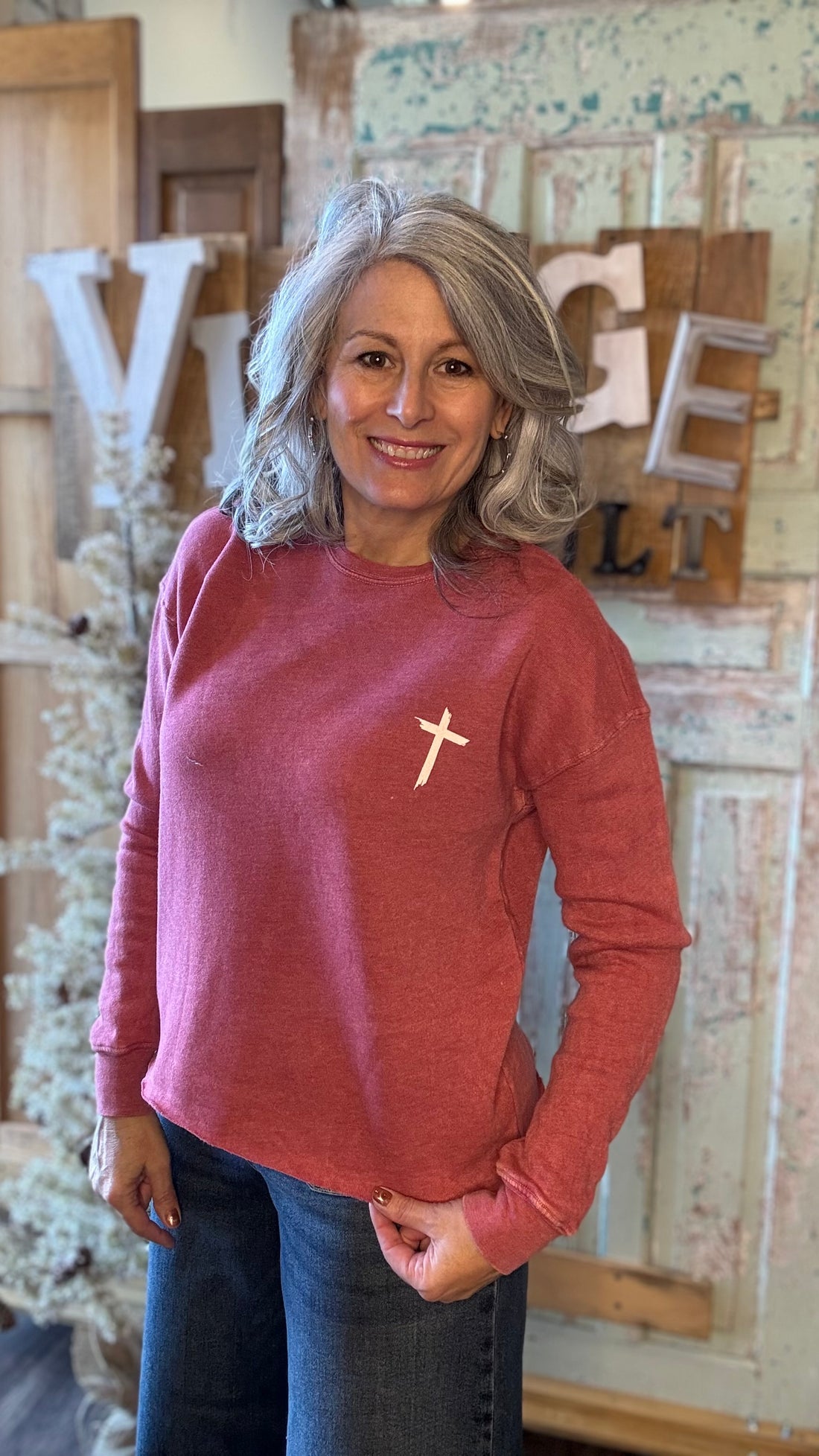 Woman wearing a red long-sleeve shirt with a cross design, standing in front of rustic wooden letters.
