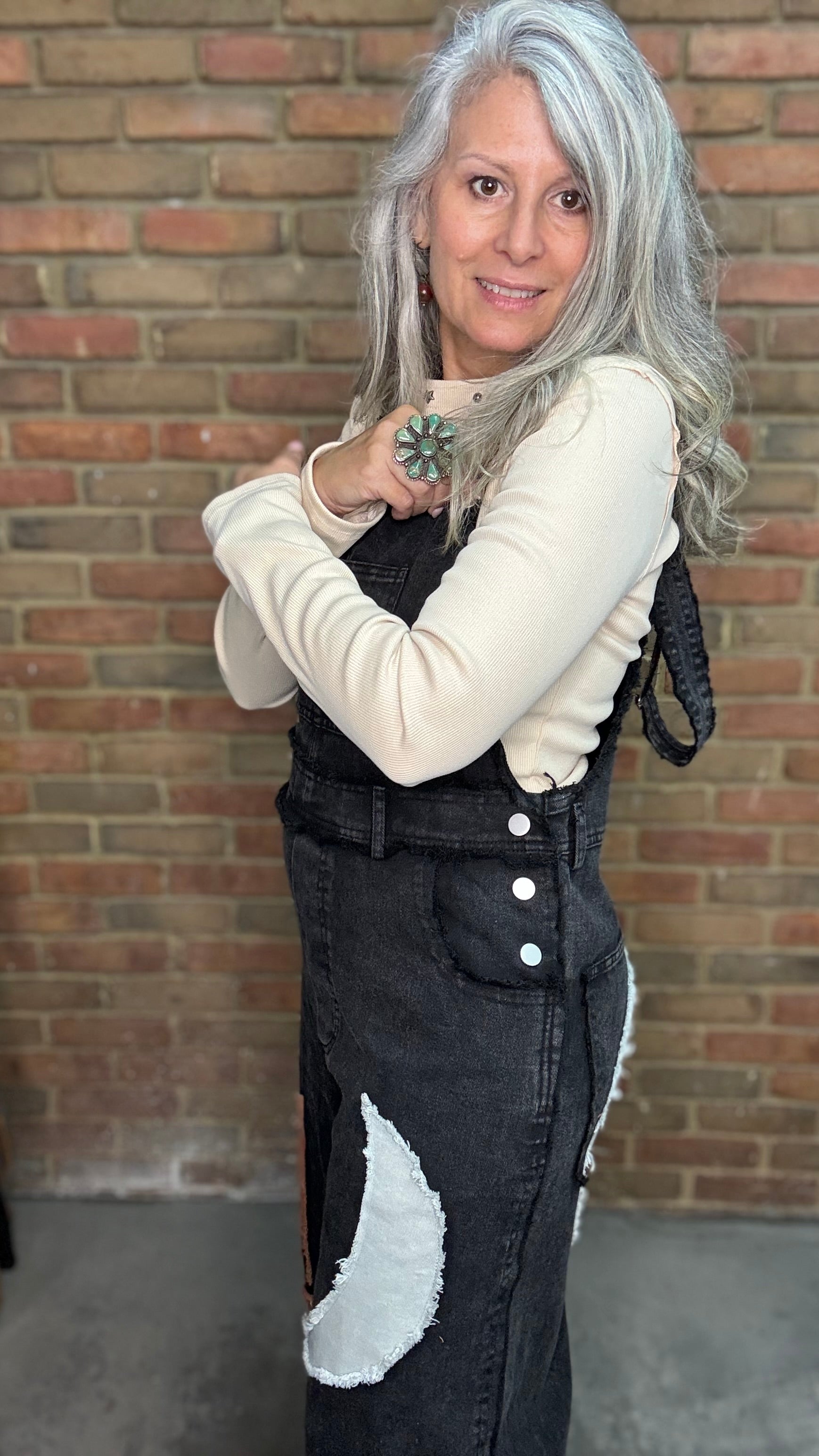 A woman with long silver-gray hair stands in front of a brick wall wearing a cream long-sleeve top layered under black denim overalls with a white patch detail on the leg.