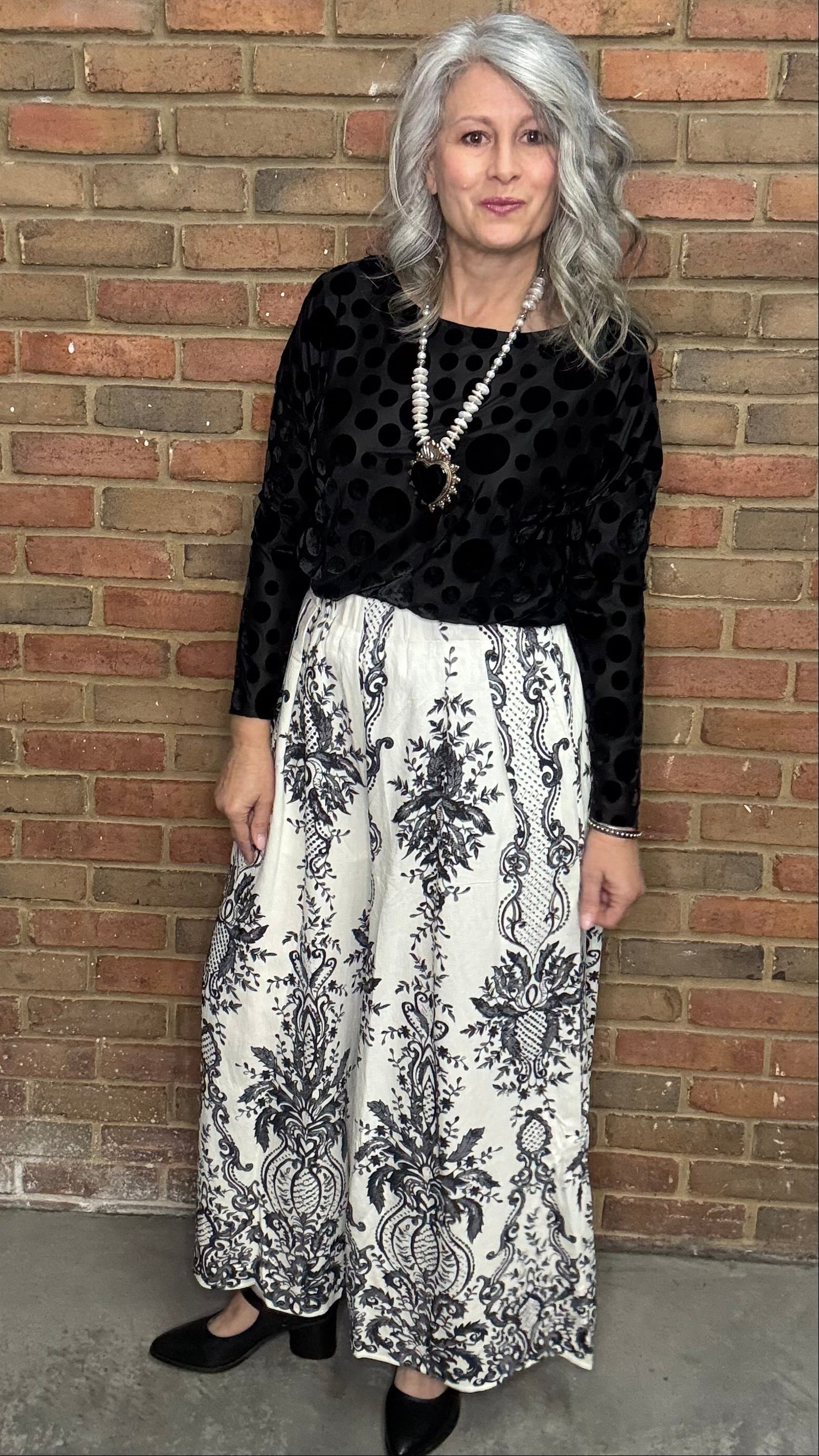 Woman wearing Black Velvet Burnout Top, Floral Print Wide Leg Pant against a brick wall. 