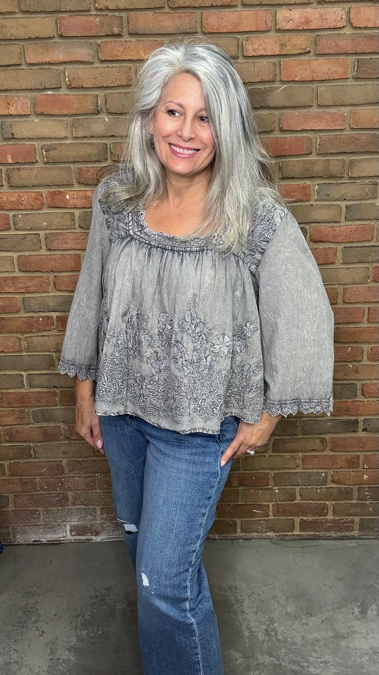 Woman with gray hair wearing the Vintage Washed Floral Lace Top a  light gray blouse and blue jeans standing against a brick wall.