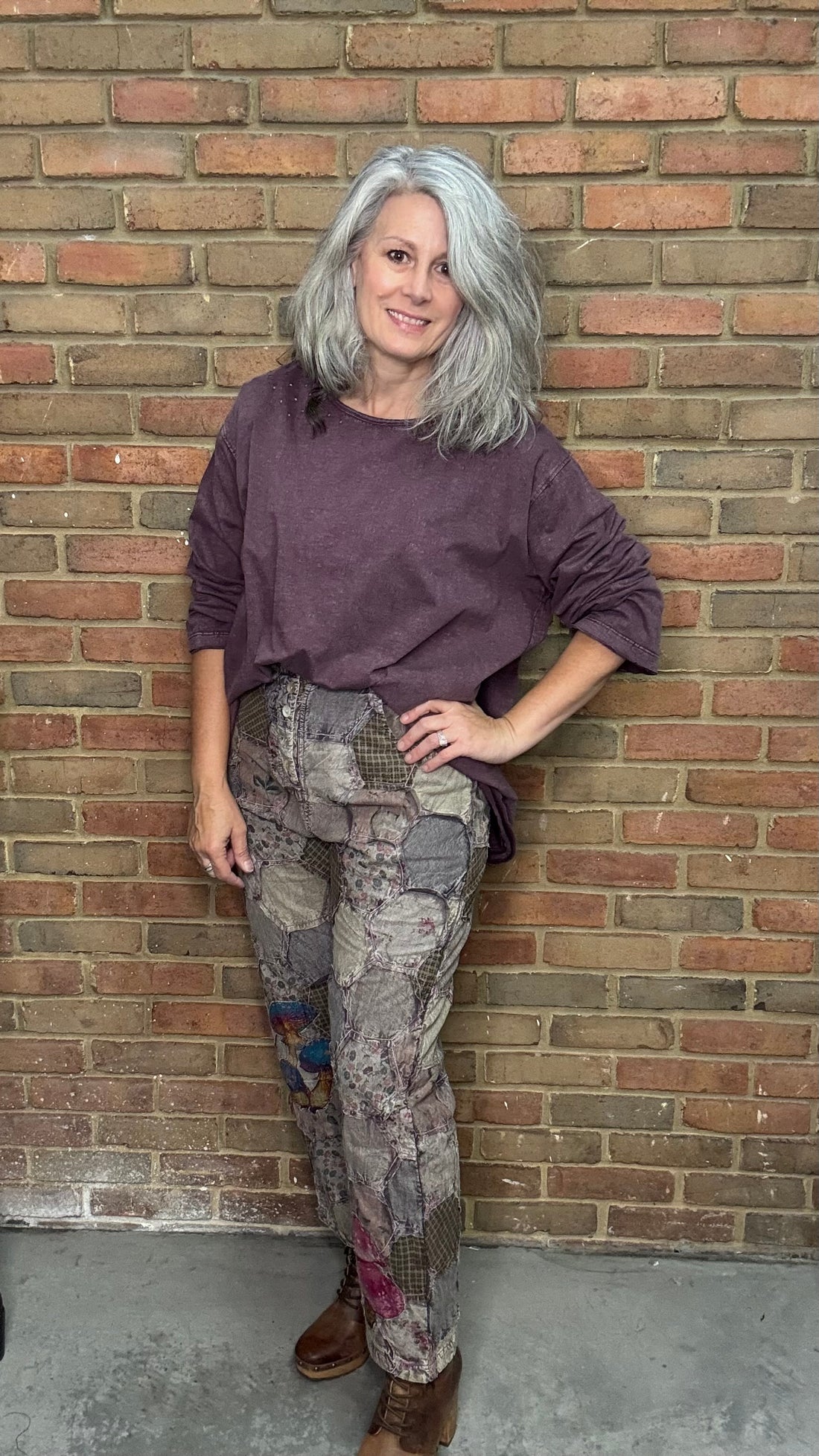 Woman leaning against a brick wall wearing a purple top and patterned pants.