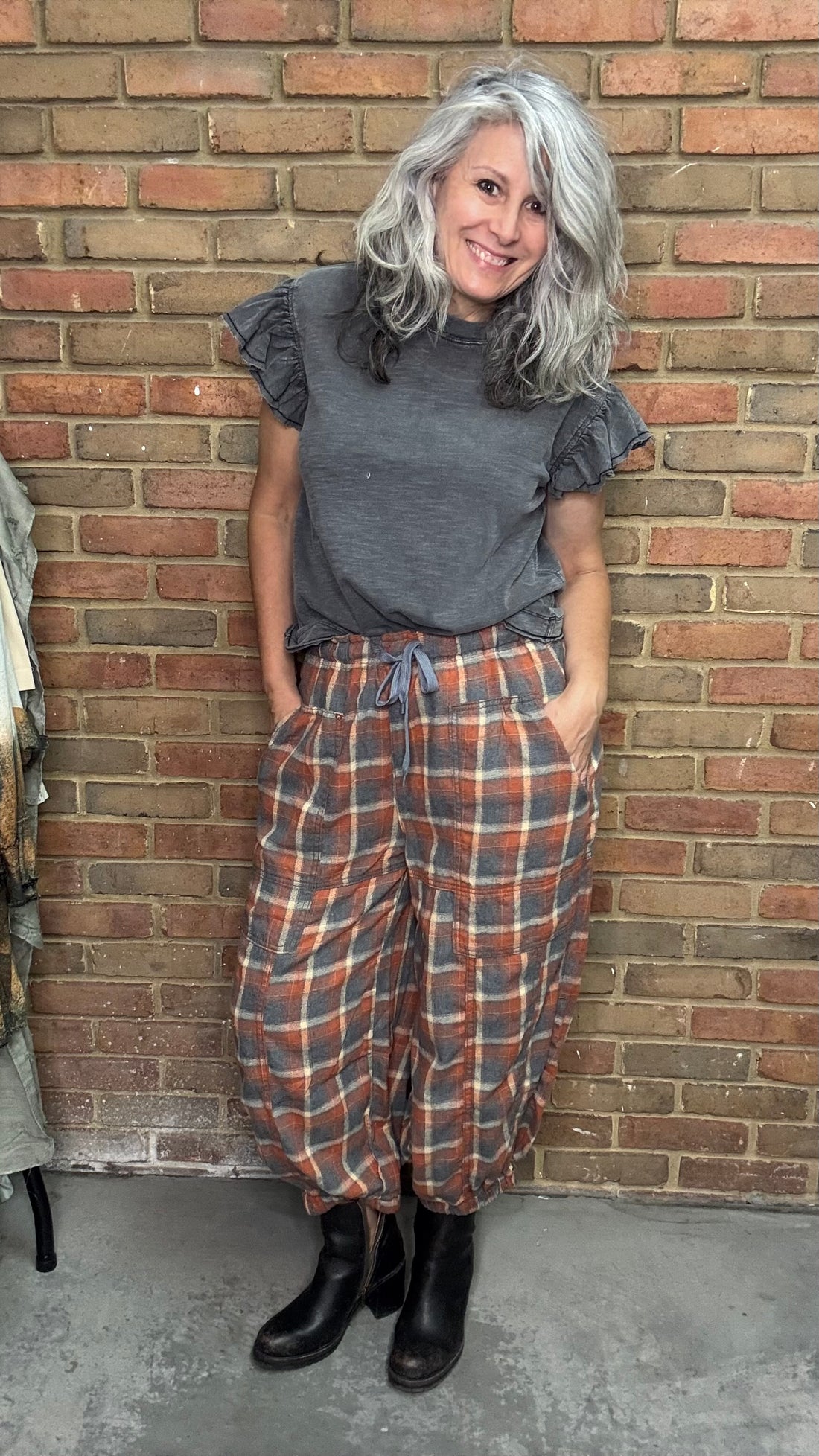 Woman wearing a gray ruffled top and plaid pants standing against a brick wall.