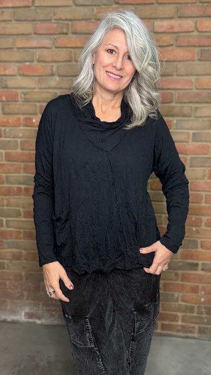 Woman wearing a black long-sleeve top and dark pants standing against a brick wall.