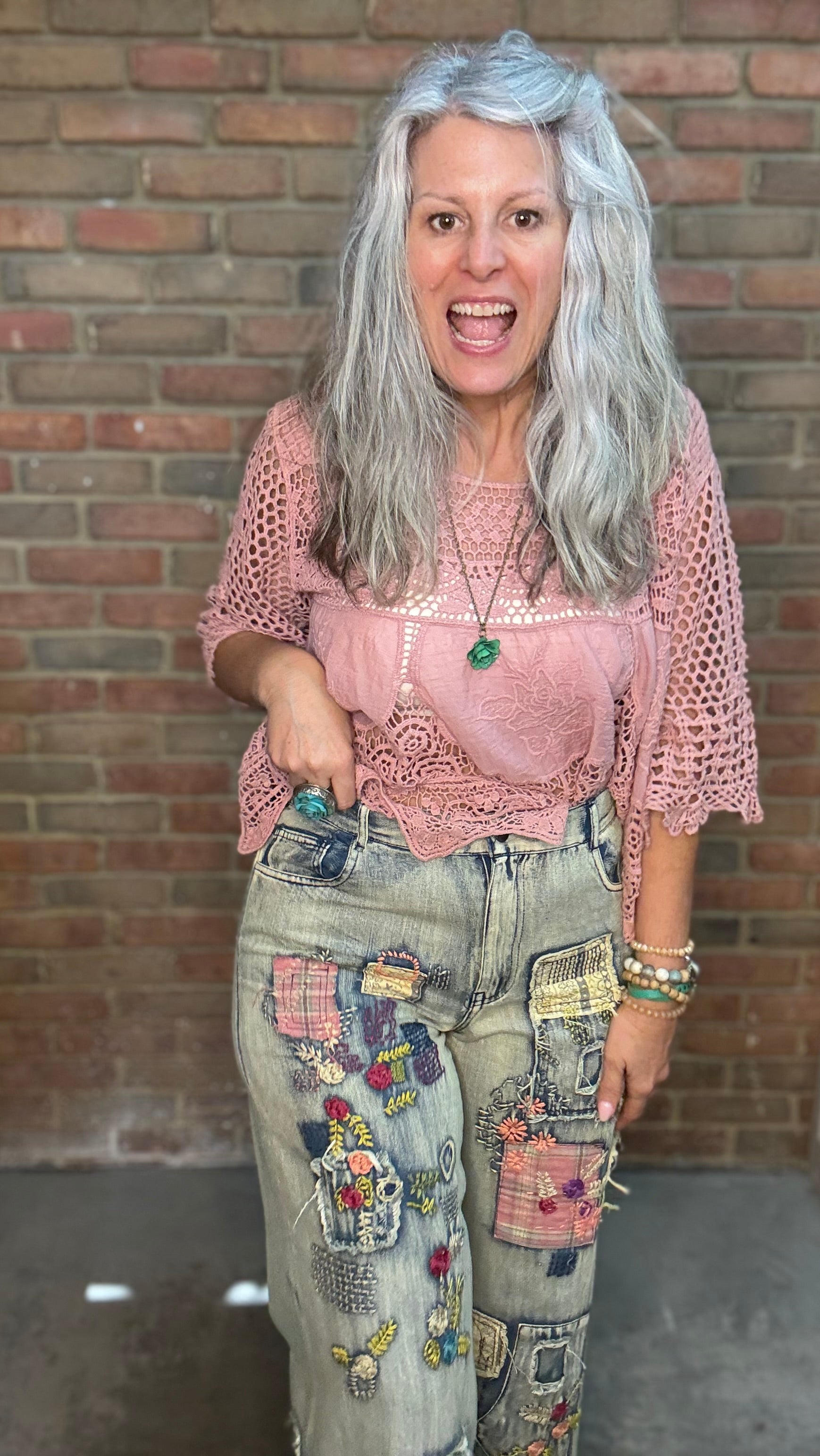 A woman with long gray hair stands in front of a brick wall wearing a pink crochet top, light-wash distressed jeans with colorful patchwork pockets, and multiple bracelets.