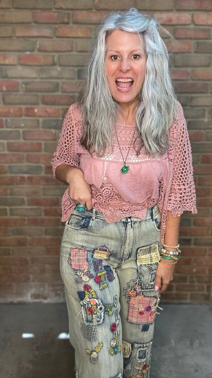 A woman with long gray hair stands in front of a brick wall wearing a pink crochet top, light-wash distressed jeans with colorful patchwork pockets, and multiple bracelets.