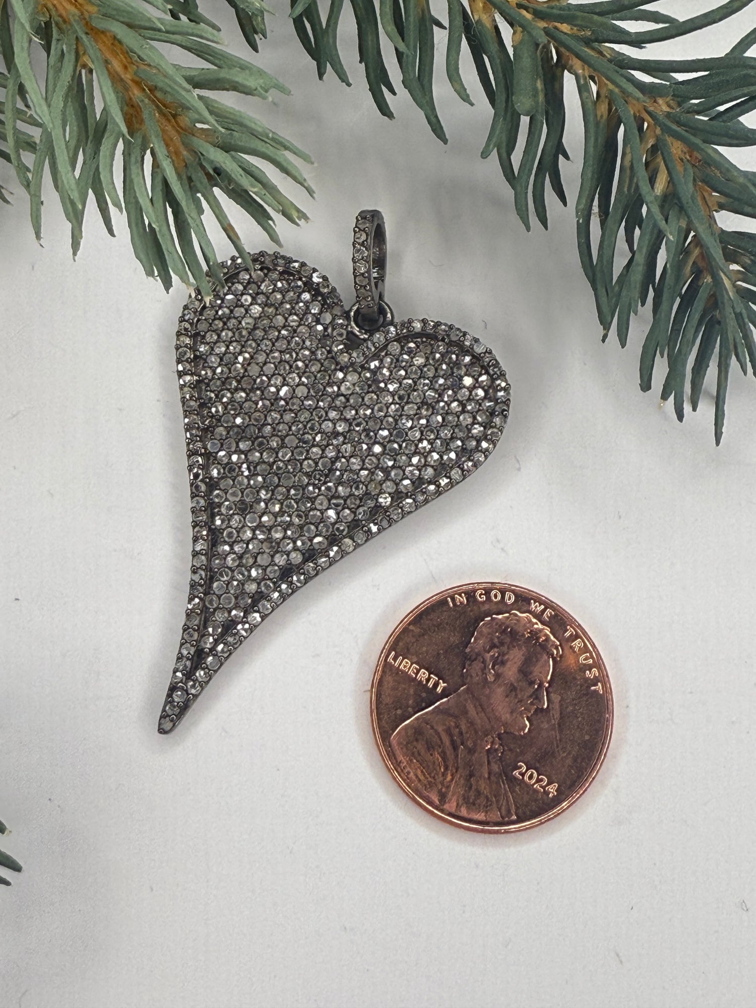 Heart-shaped pendant with pave diamond stones next to a penny on a light background with greenery.