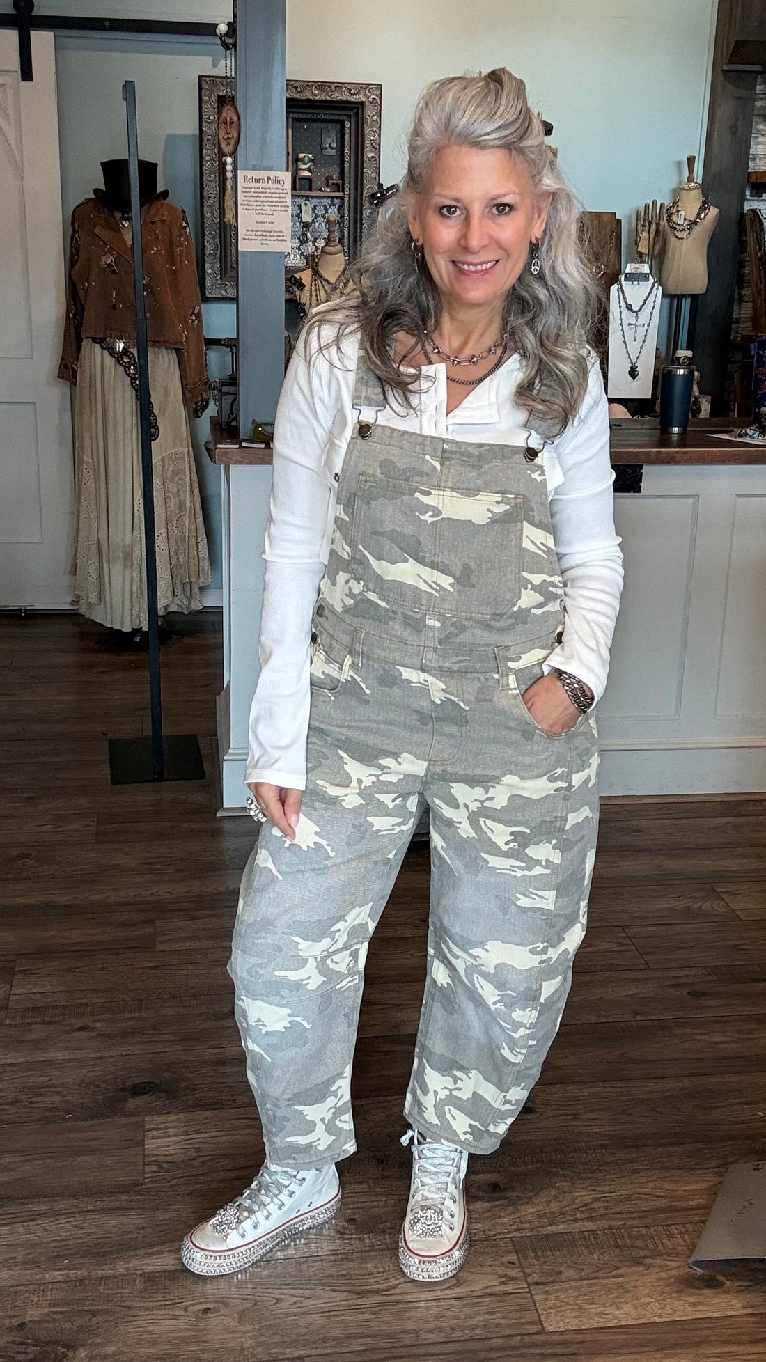 A woman with gray hair stands in a boutique wearing gray camouflage overall over a white long-sleeve shirt and white sneakers.
